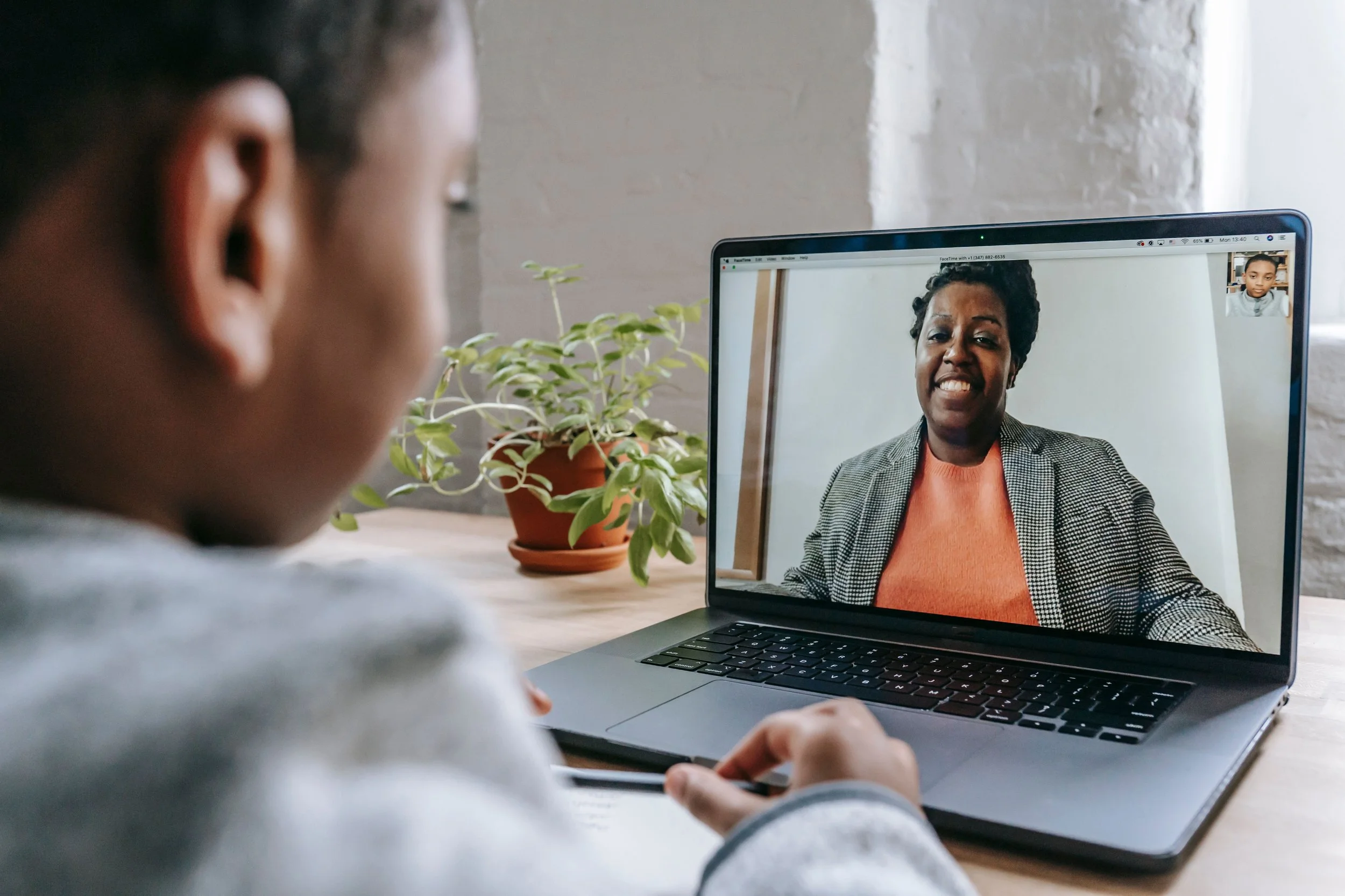 A child participating in a video call on a laptop, with a woman visible on the screen, smiling and dressed in a blazer and orange top, in a bright, cozy room with a potted plant.