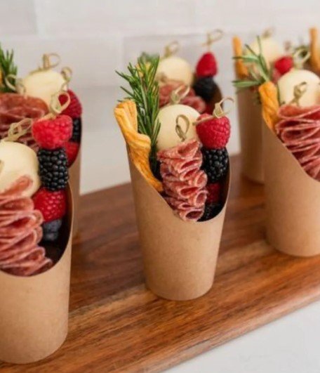 Appetizer cups with salami, cheese, raspberries, blackberries, and herbs on a wooden tray.