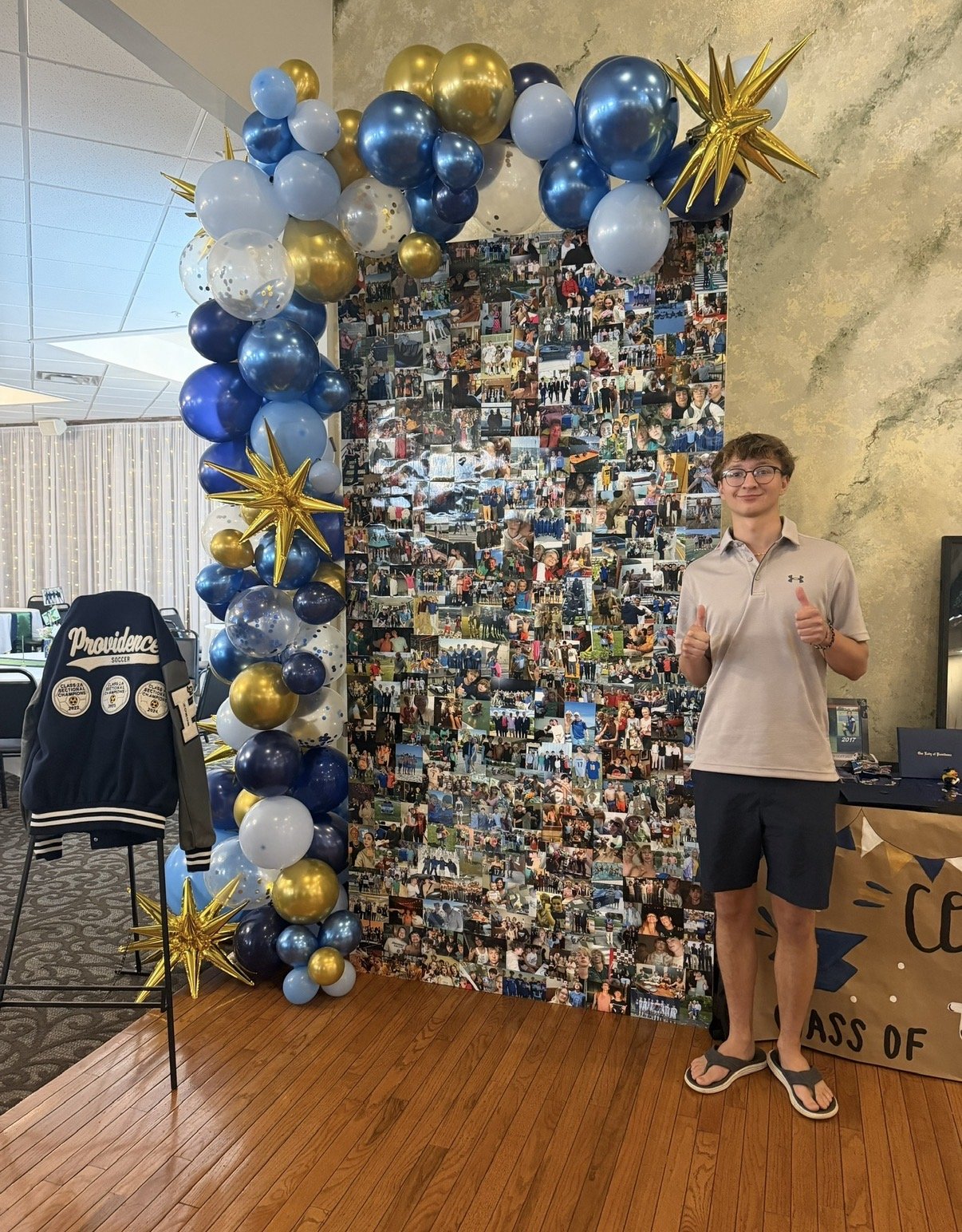 A young man stands next to a photo collage and balloon arch at an indoor event. The collage features numerous small pictures of people celebrating. The balloon arch is decorated with blue, white, gold, and navy balloons, and gold star-shaped balloons. A Providence soccer jacket hangs on a chair nearby, and a person is giving a thumbs-up gesture, smiling.