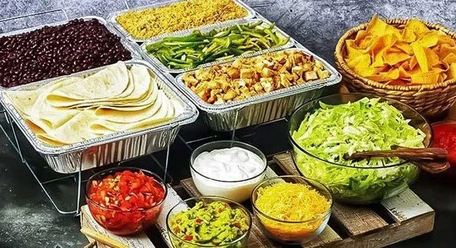 Assorted ingredients for a Mexican taco bar including black beans, shredded cheese, sliced green peppers, cooked chicken, tortilla chips, shredded lettuce, diced tomatoes, guacamole, sour cream, shredded yellow cheese, and salsa in bowls.