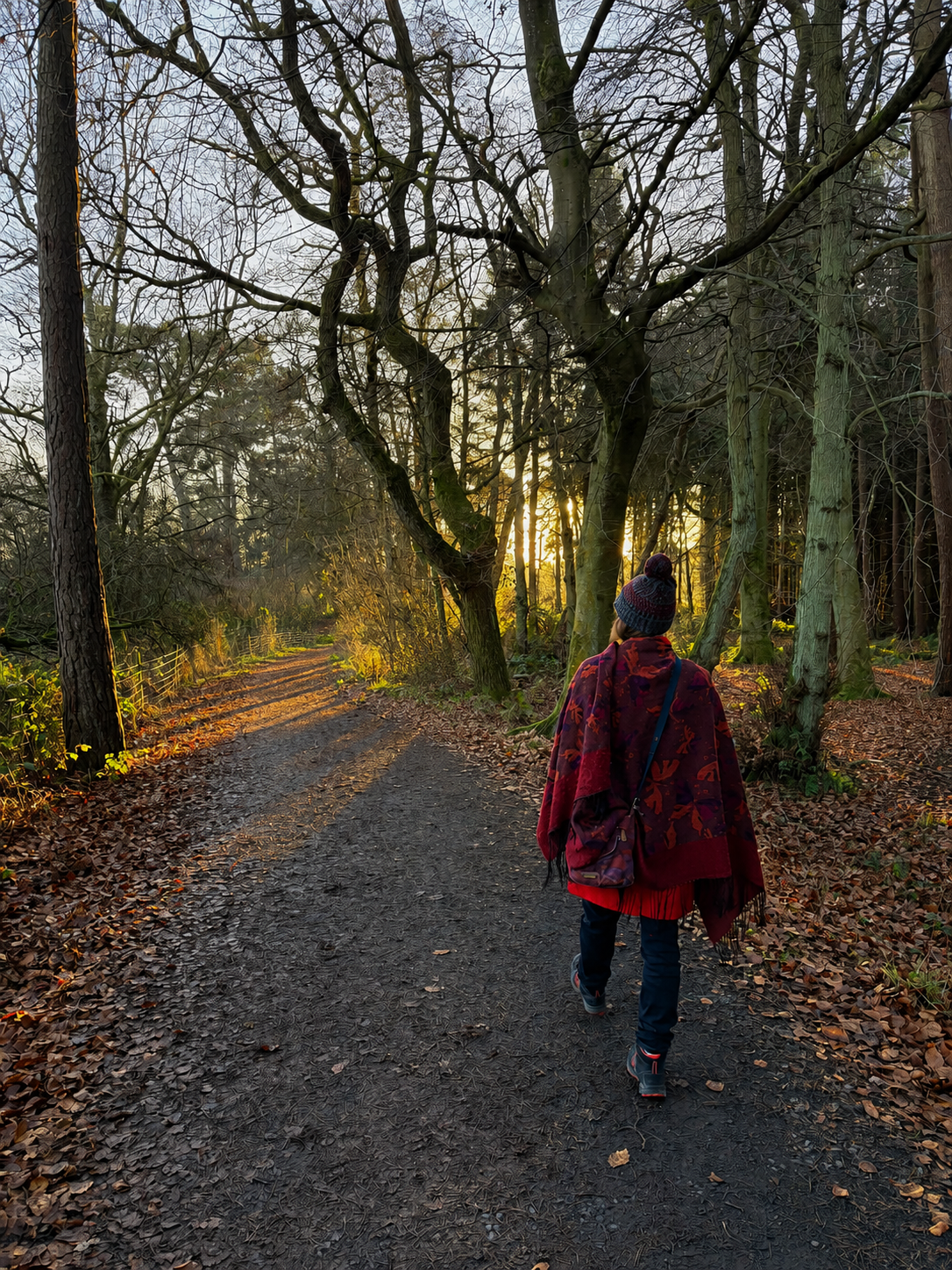 Person walking on a dirt path in a wooded forest during sunset, wearing a red patterned shawl, hat, and carrying a backpack.