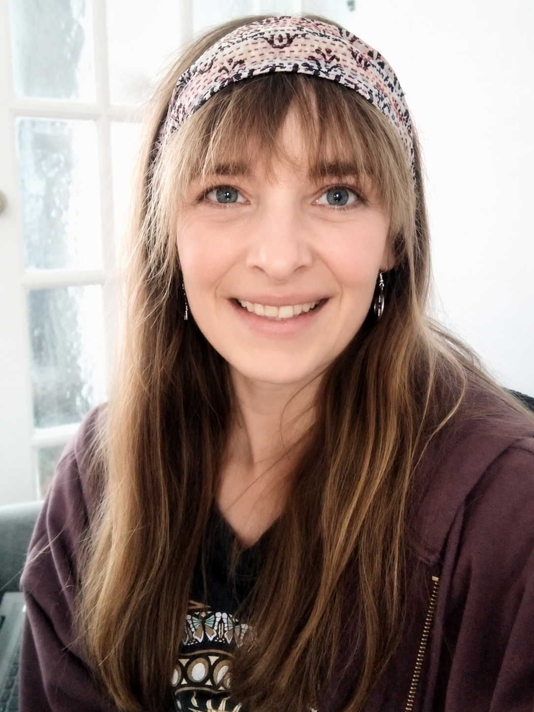 A young woman with long, light brown hair, wearing a patterned headband and earrings, smiling at the camera indoors near a window.