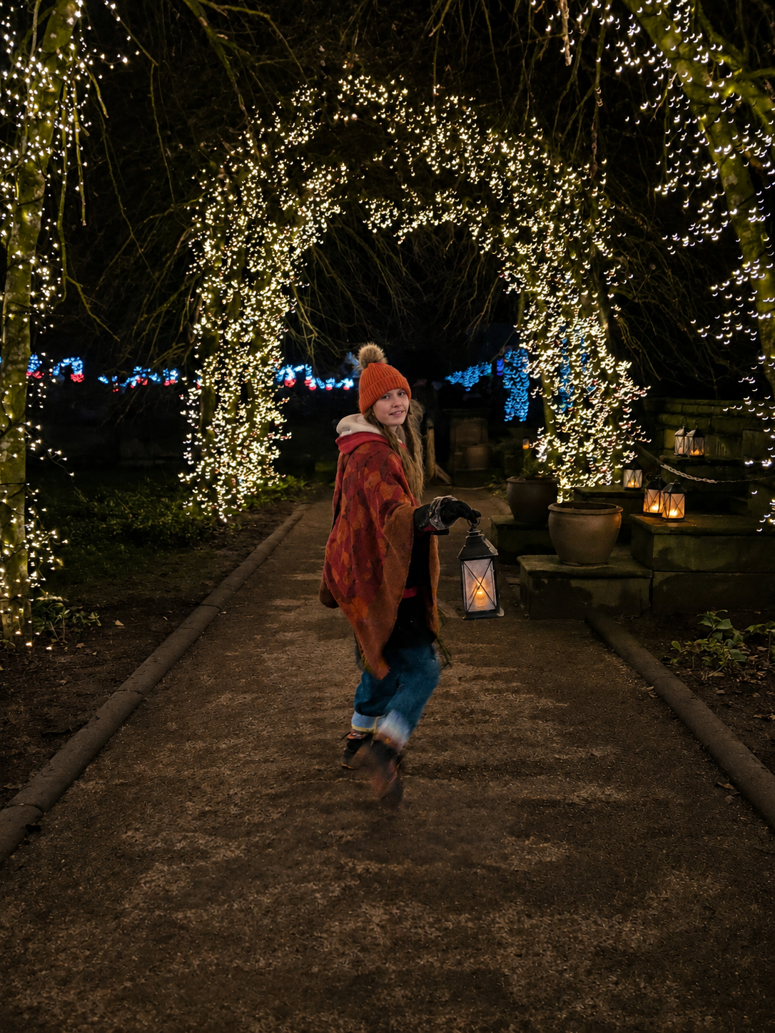 A young woman wearing a red knit hat, a colorful shawl, and winter clothing, holding a lantern, walking through a pathway decorated with holiday string lights and lanterns at night.
