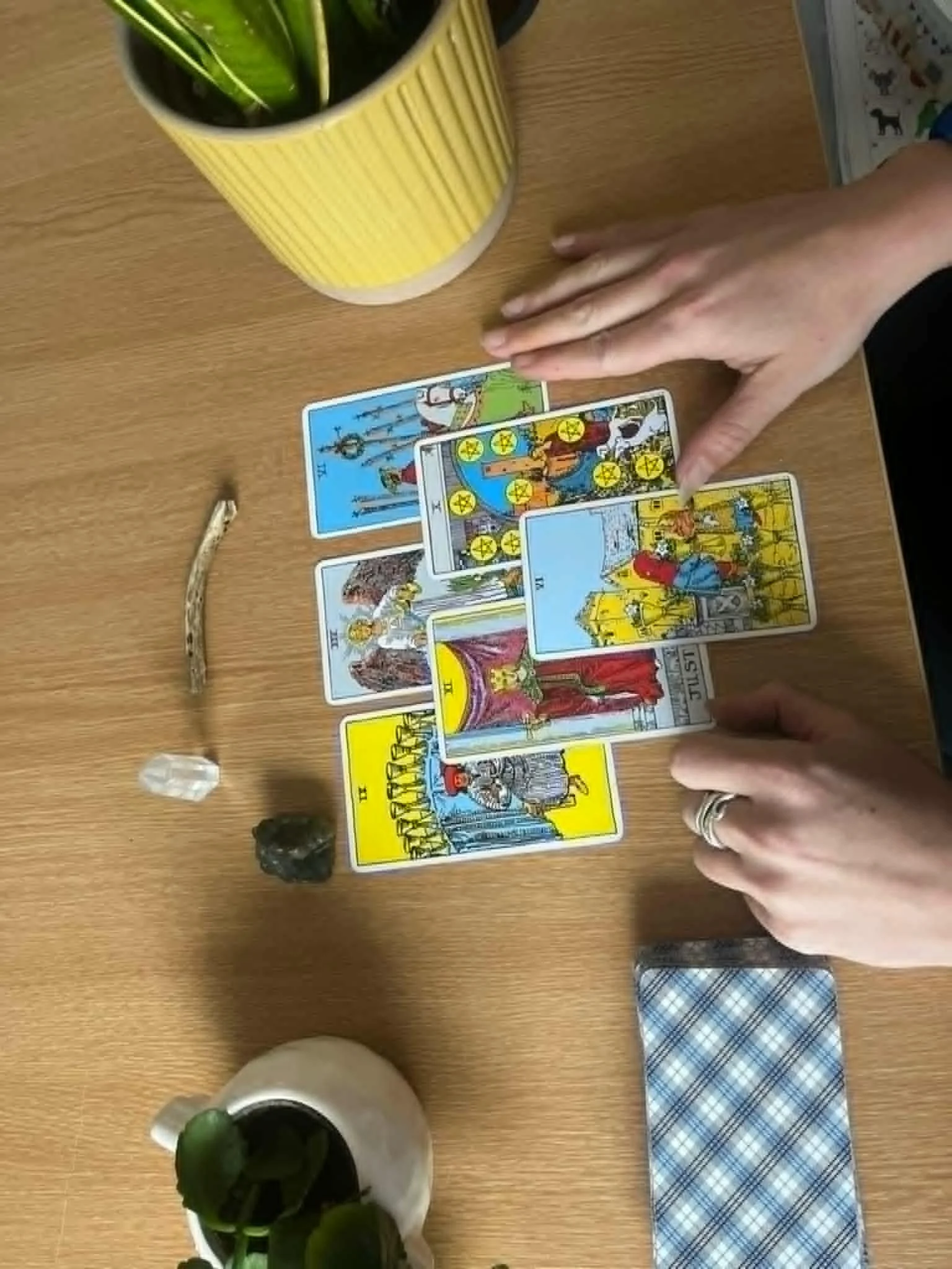 A person's hand is seen over a tarot card spread on a wooden table, with tarot cards showing various imagery laid out. There are potted plants, a yellow striped cup, and some other items on the table.