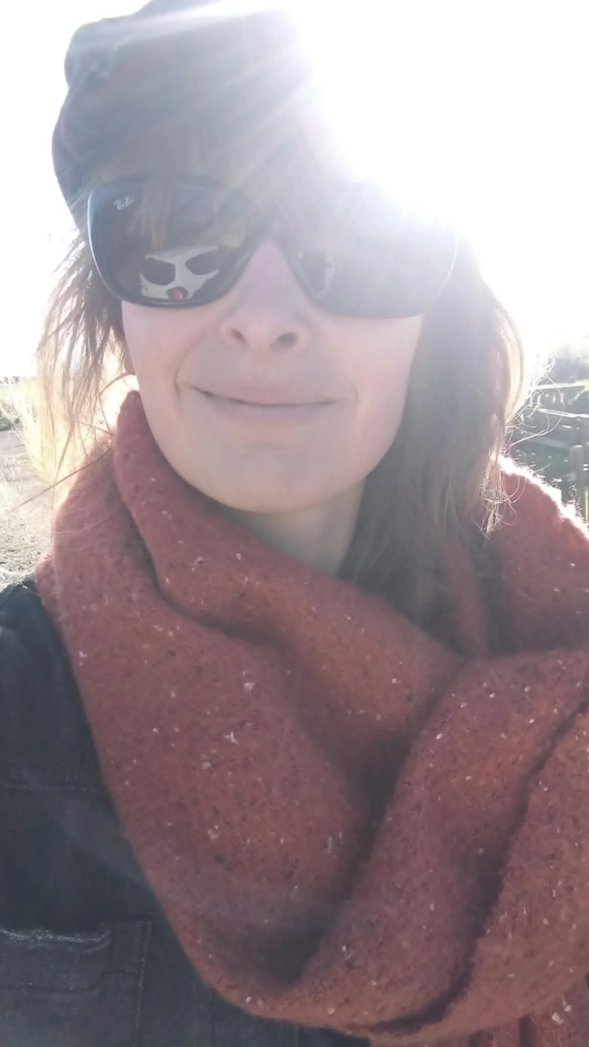Person wearing sunglasses and a beanie, smiling in bright sunlight, wrapped in a rust-colored scarf.
