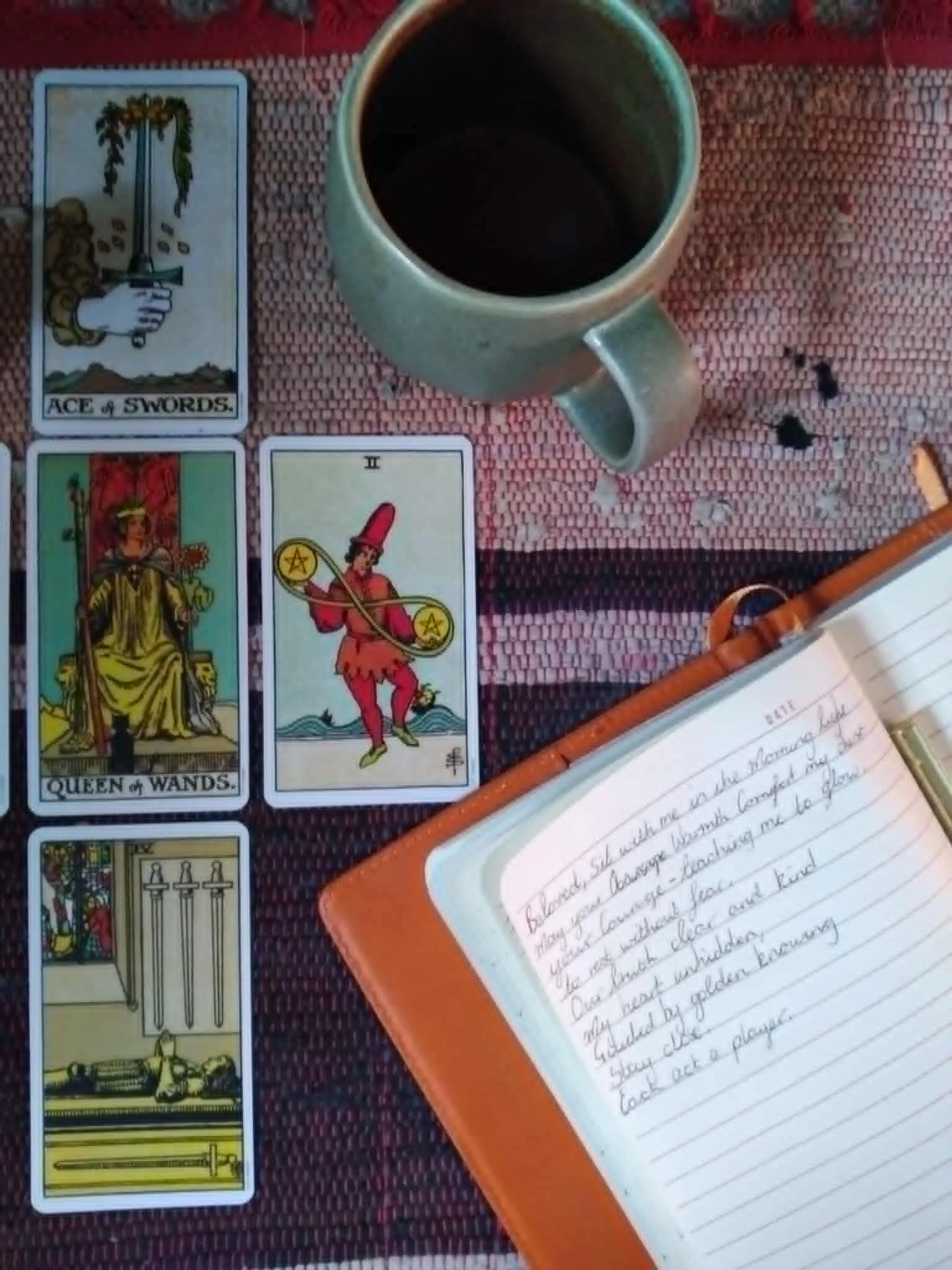 A cup of black coffee, three tarot cards, and a handwritten journal on a table with a woven, red and black patterned tablecloth.