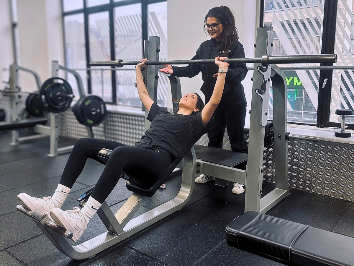 A woman spotting another woman lifting weights on a weight bench in a gym.