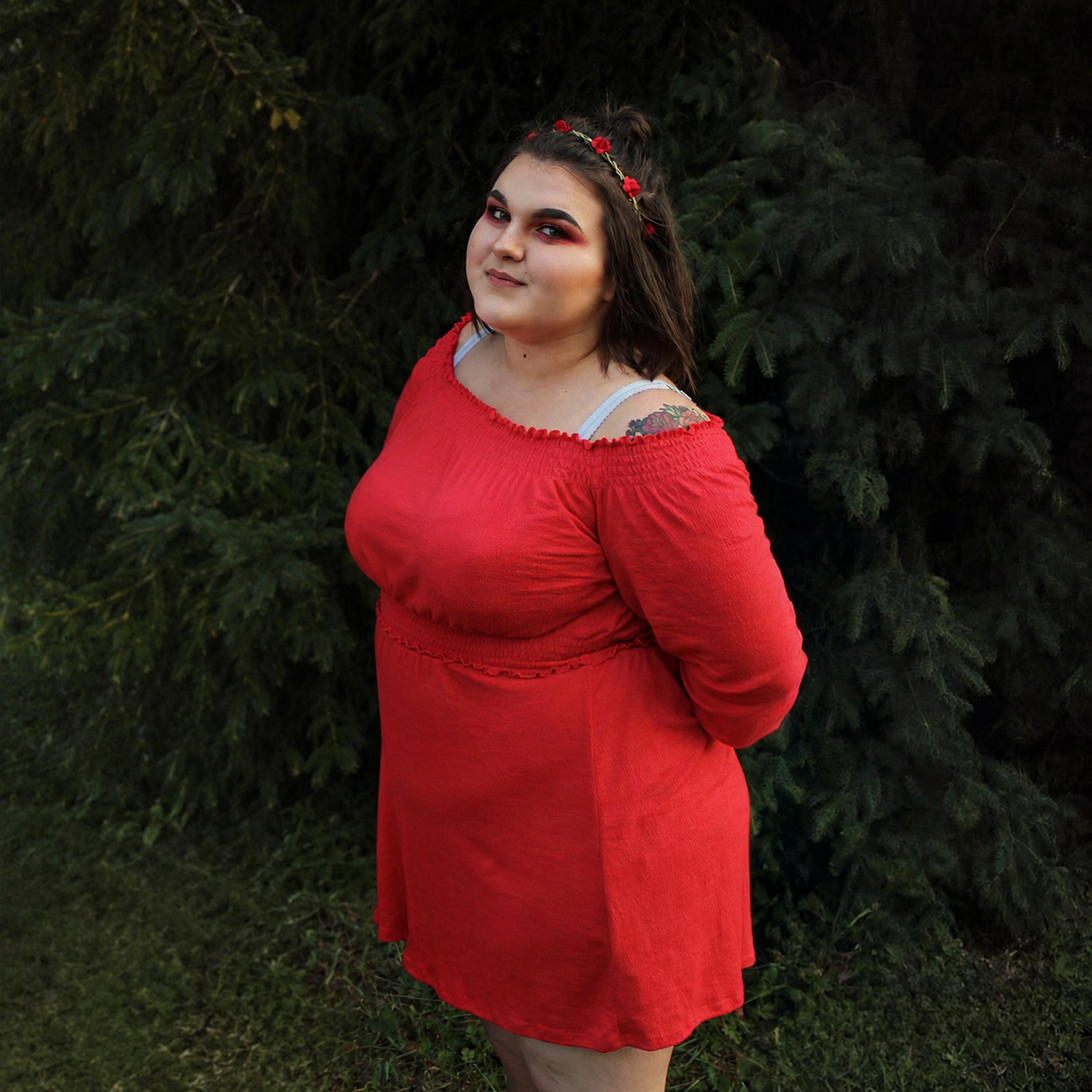 A woman in a red off-shoulder dress with a white strap visible underneath, standing outdoors in front of a lush green evergreen tree, with a flower crown on her head and makeup highlighting her eyes.