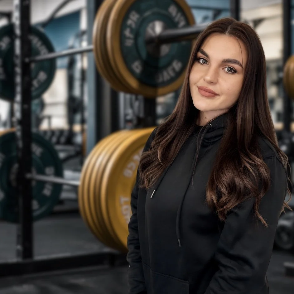 Young woman with long brown hair and a black hoodie in a gym with weightlifting equipment and weights in the background.
