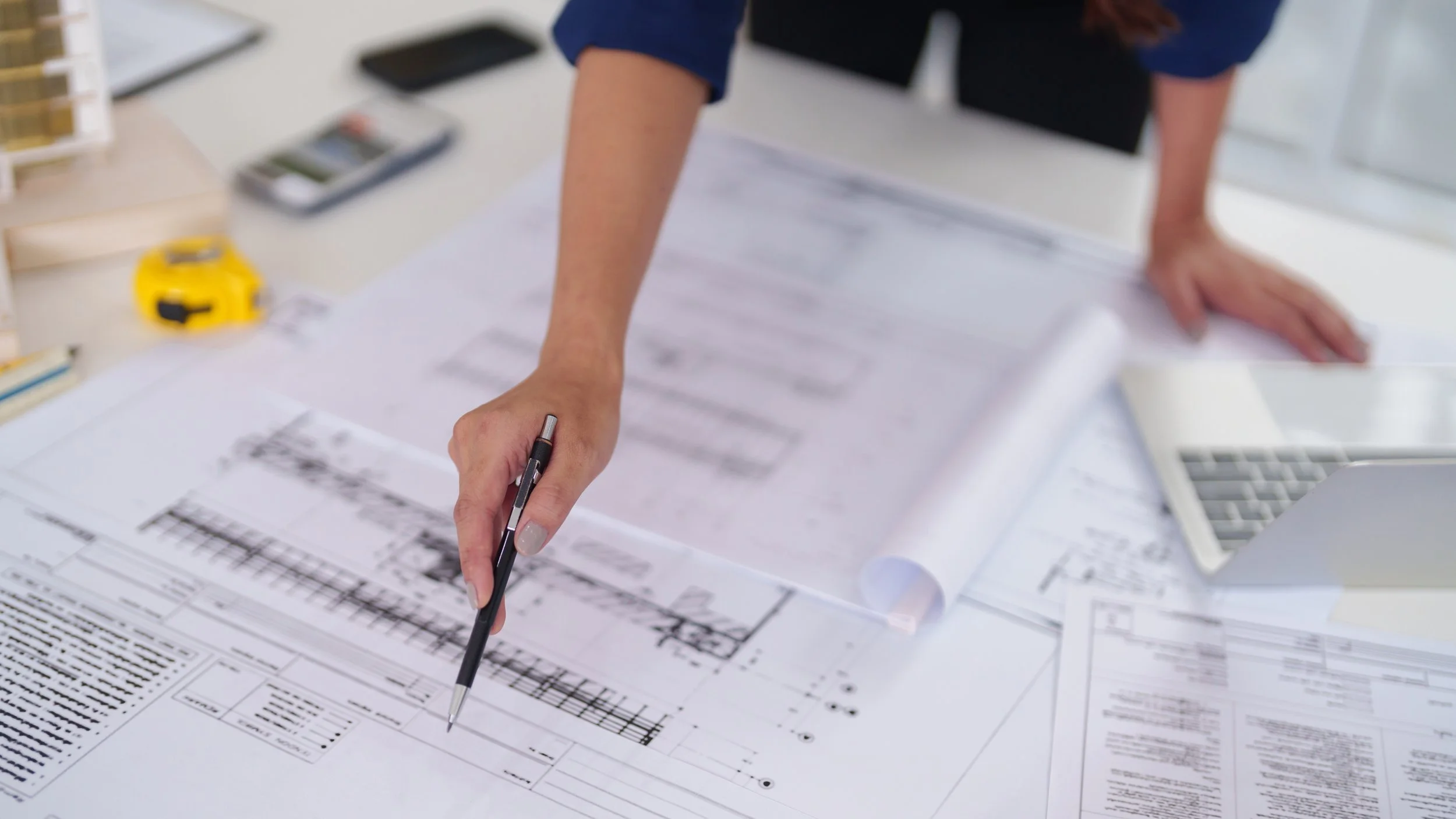 Person reviewing architectural blueprints on a table with various office supplies.