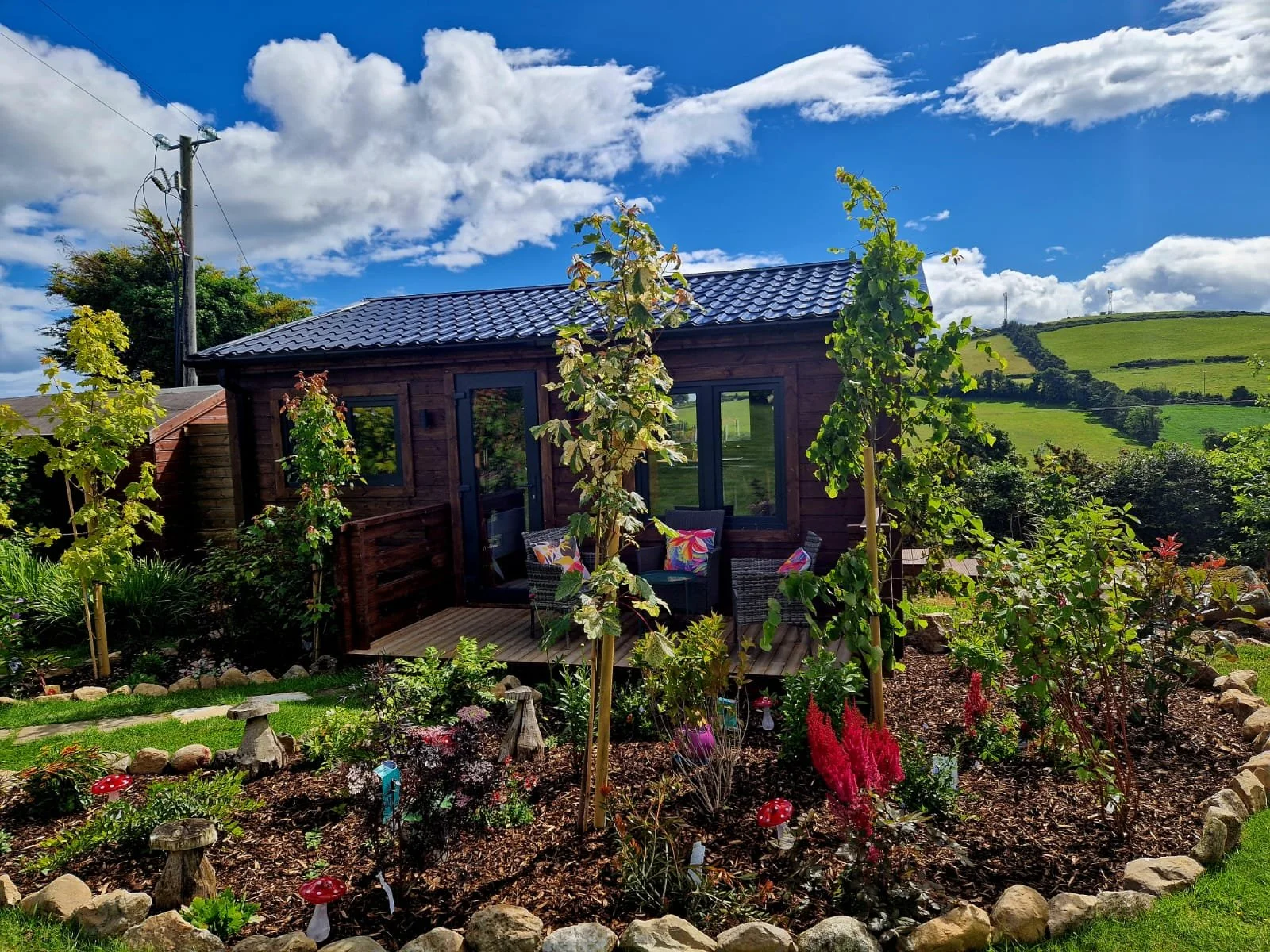Small wooden garden house with a blue tiled roof, surrounded by lush greenery, colorful flowers, and a garden sitting area under a bright blue sky with clouds.