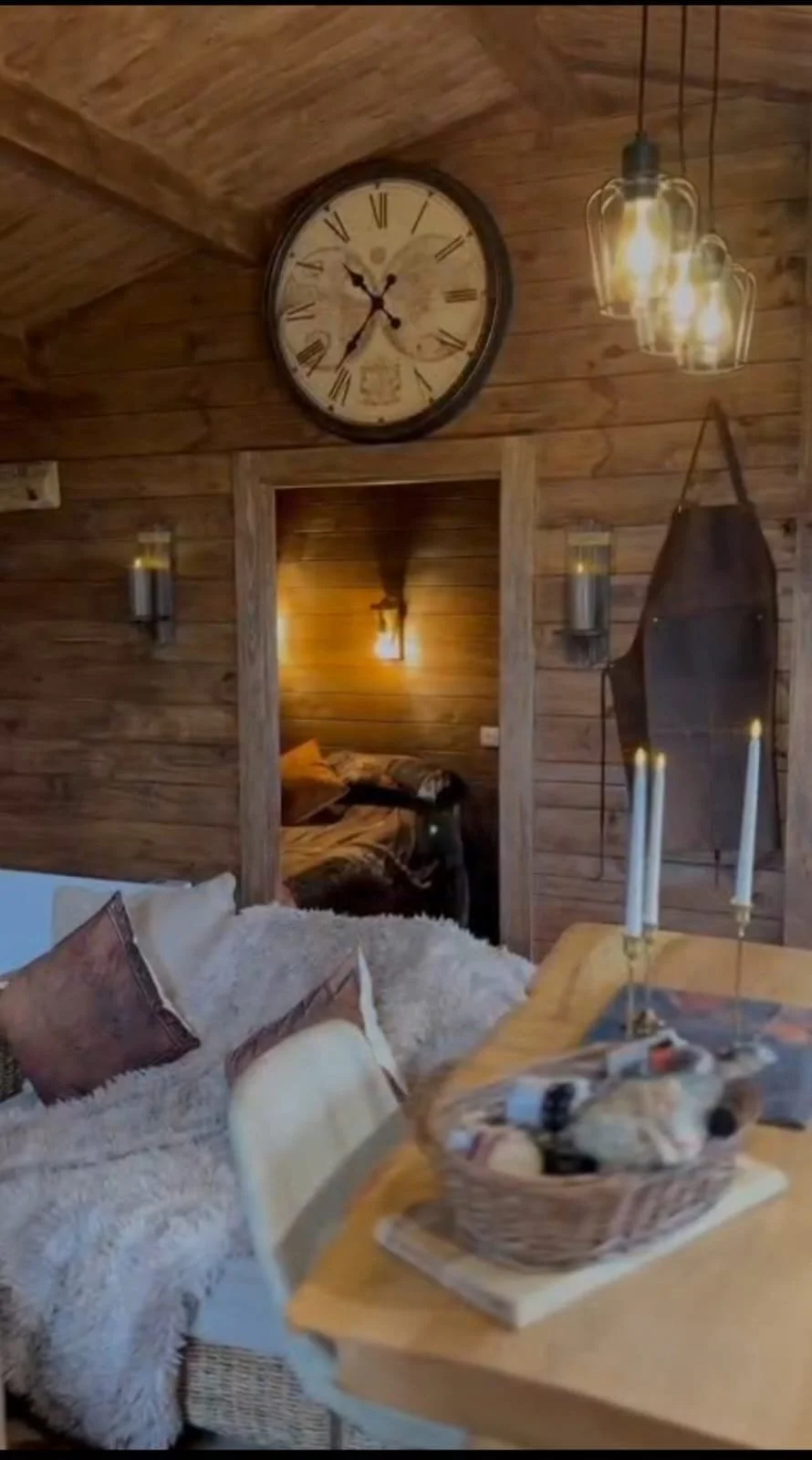 Wood-paneled living room with a large clock on the wall, hanging lights, a cozy bed visible through a doorway, a cushioned sofa with pillows, and a basket of logs on a table.