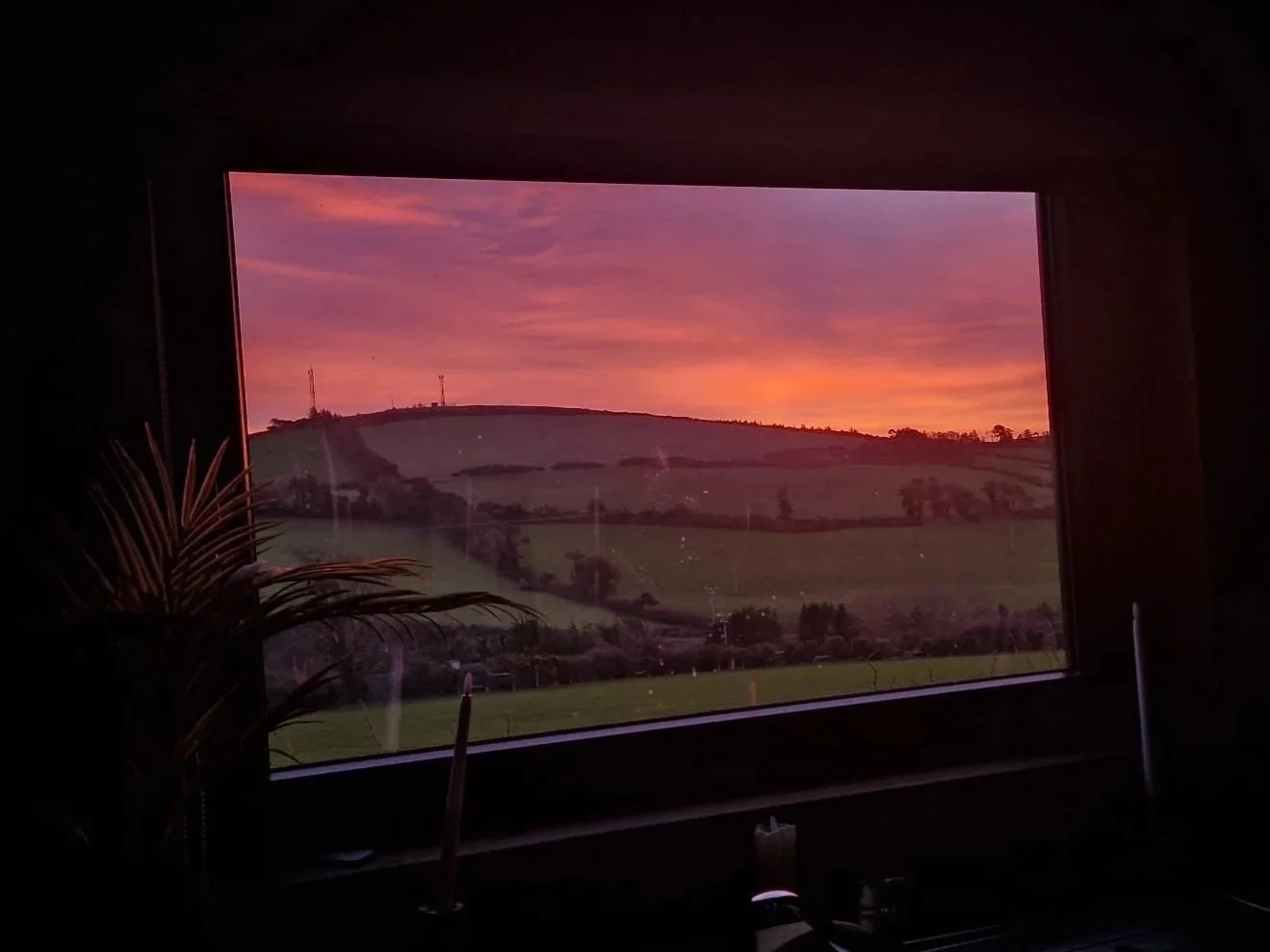 View of a colorful sunset sky over grassy hills seen through a window indoors.