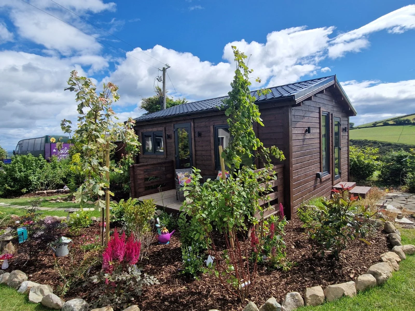 Small wooden house surrounded by a garden with colorful plants, flowers, and trees under a blue sky with white clouds.
