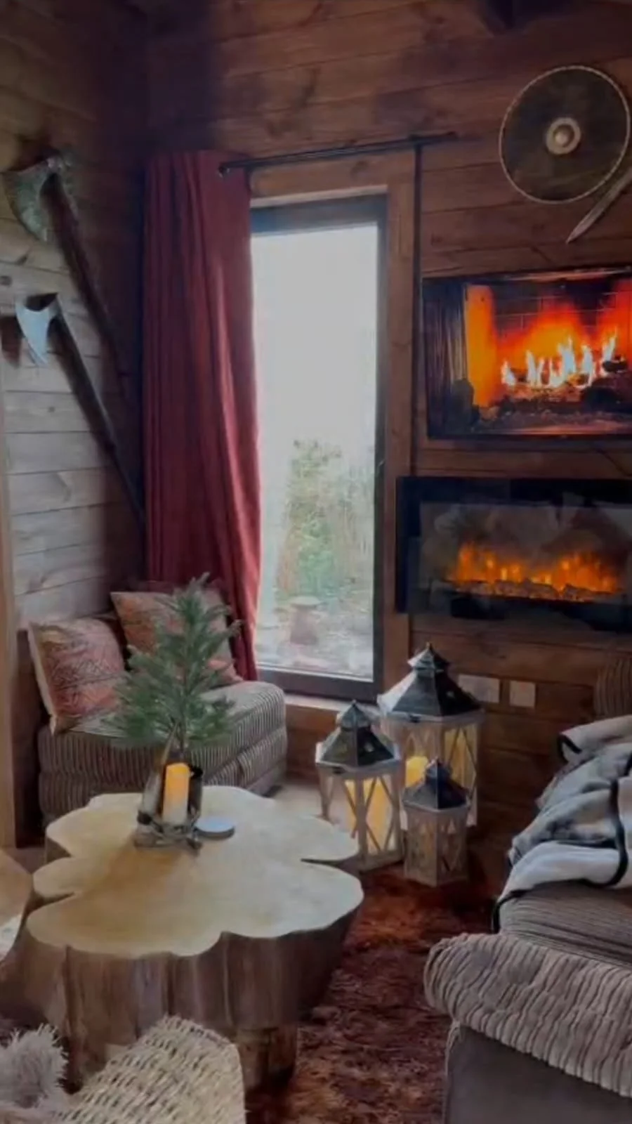 Cozy living room with wood-paneled walls, a large window with red curtains, a sofa with striped cushions, a tree-shaped decor on a wooden table, lanterns on the floor, and two electric fireplaces on the wall, with a mounted fan and axe on the walls.