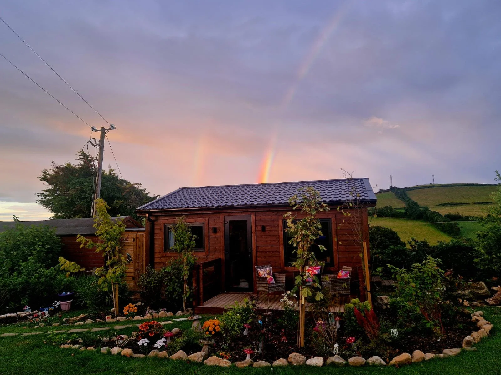 A cozy wooden garden shed with a blue tiled roof, surrounded by planted trees and flowers, with a sunset sky and a rainbow in the background.