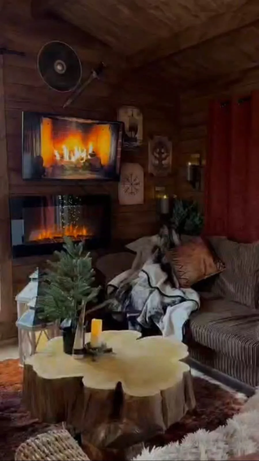 Cozy living room with wood-paneled walls, a fireplace, a TV displaying a fireplace scene, a small Christmas tree, a couch with pillows, a blanket, and decorative wall art.