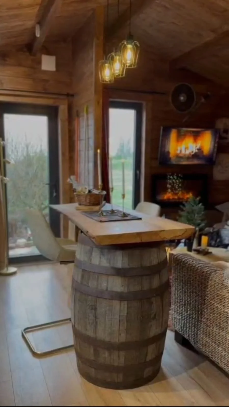 Interior of a rustic home with wood-paneled walls and ceiling, featuring a round wooden table made from a barrel, a small Christmas tree, a fireplace with a TV above, a chandelier hanging from the ceiling, and sliding glass doors with view outside.