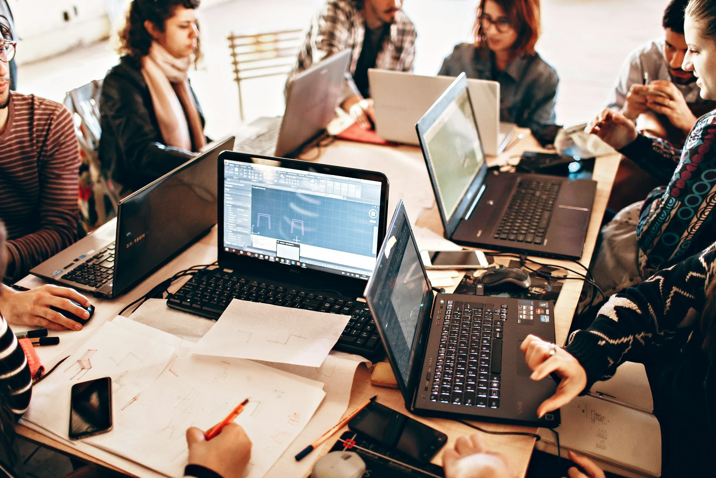 People gathered around a table working on laptops, with papers, notebooks, and writing utensils scattered across the table.