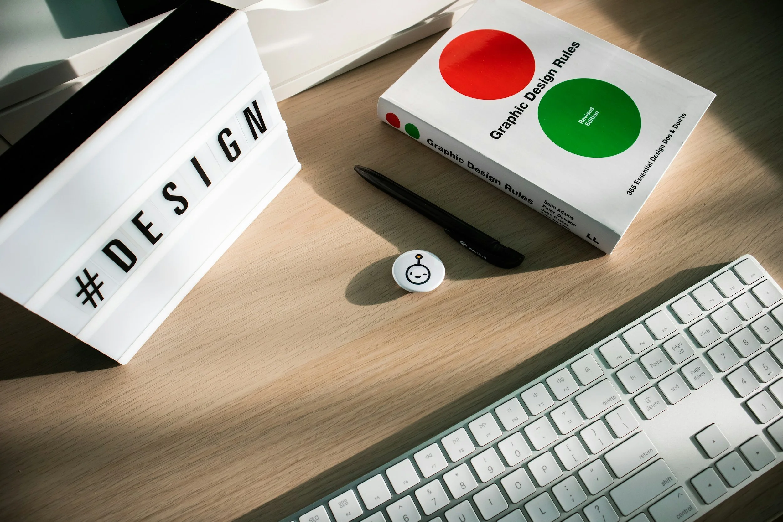 A desk with a white keyboard, a photograph of a lightbox displaying the word #DESIGN, a book titled 'Graphic Design Rules Revision Edition,' a black pen, a round clip with a smiling face, and a lightbox. The lightbox is illuminated, casting shadows on the desk.