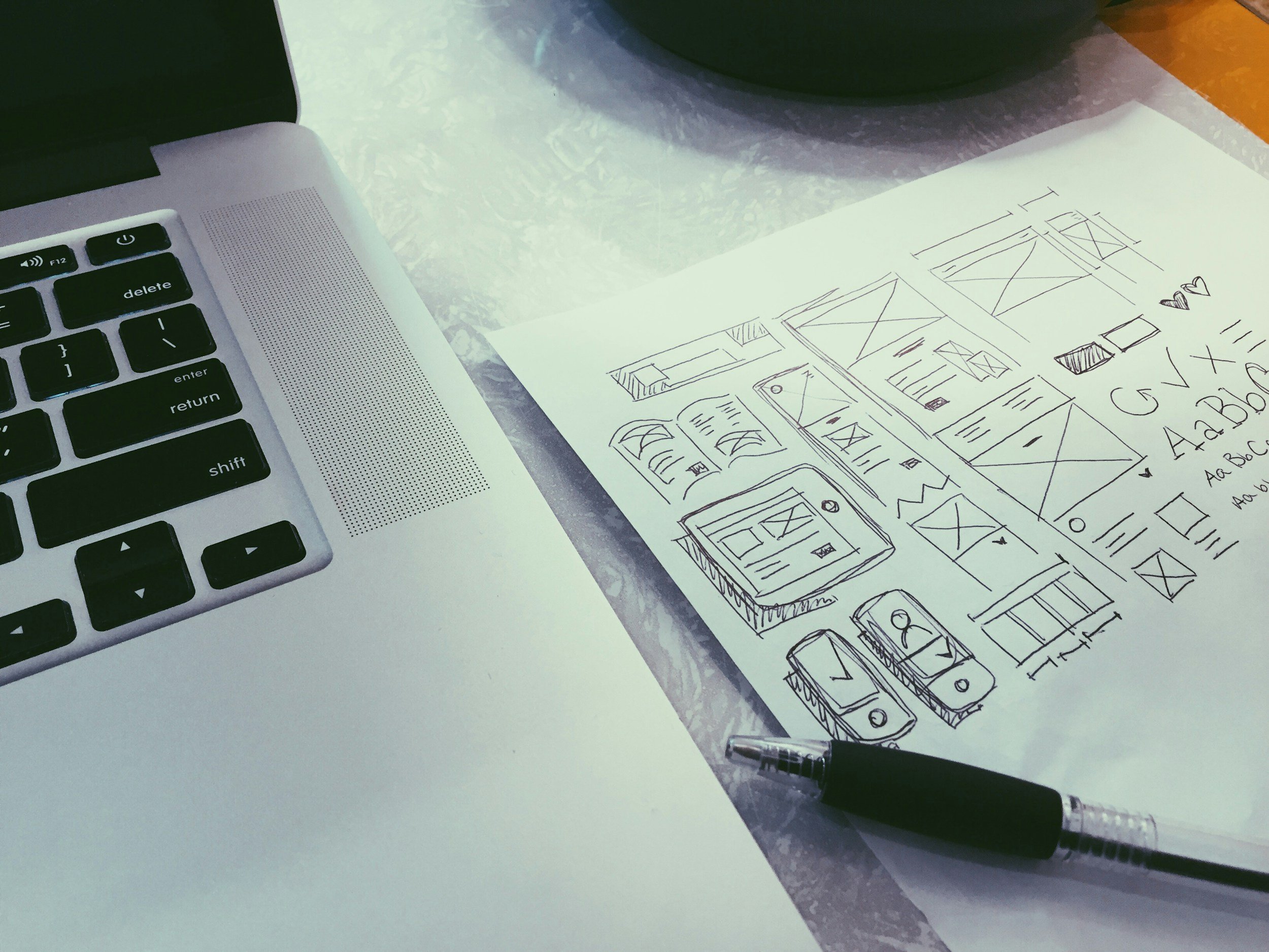 Sketches of buildings and floor plans on a sheet of paper, with a pen and a mechanical pencil nearby on a table next to a laptop.