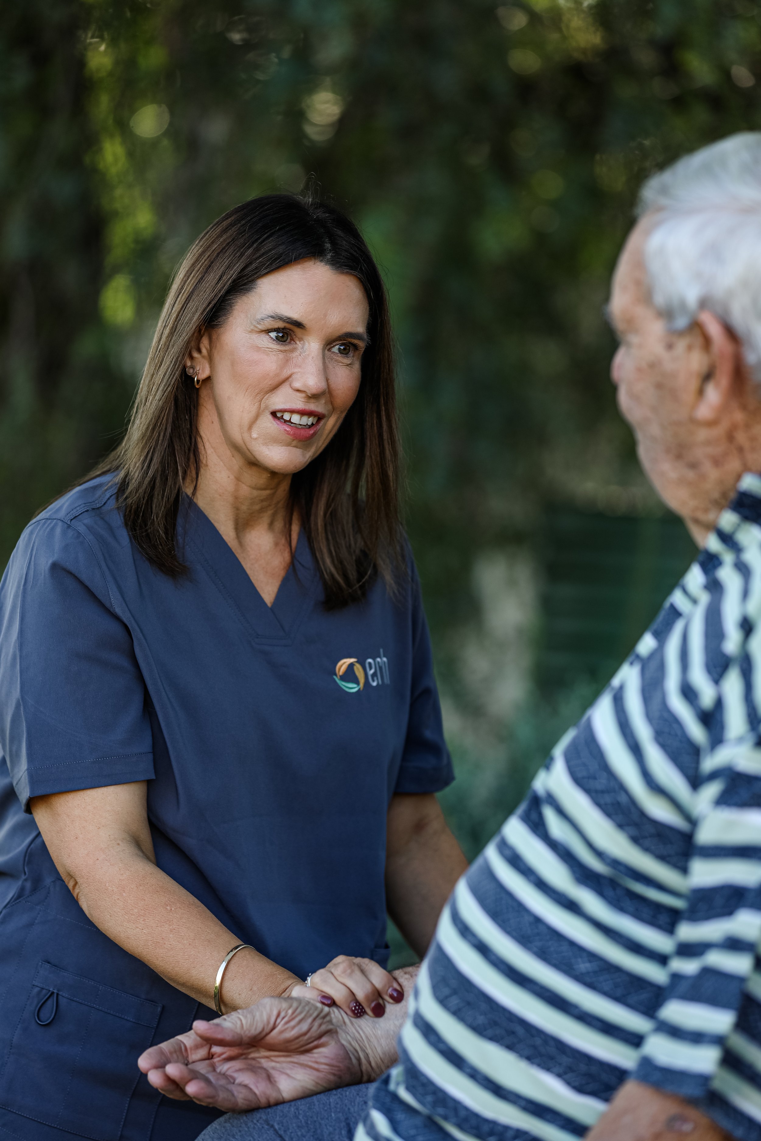 A nurse from Echuca Regional Health in a navy blue uniform completing a health check on an elderly person, who has gray hair and is wearing a striped shirt, outdoors with a blurred green background.