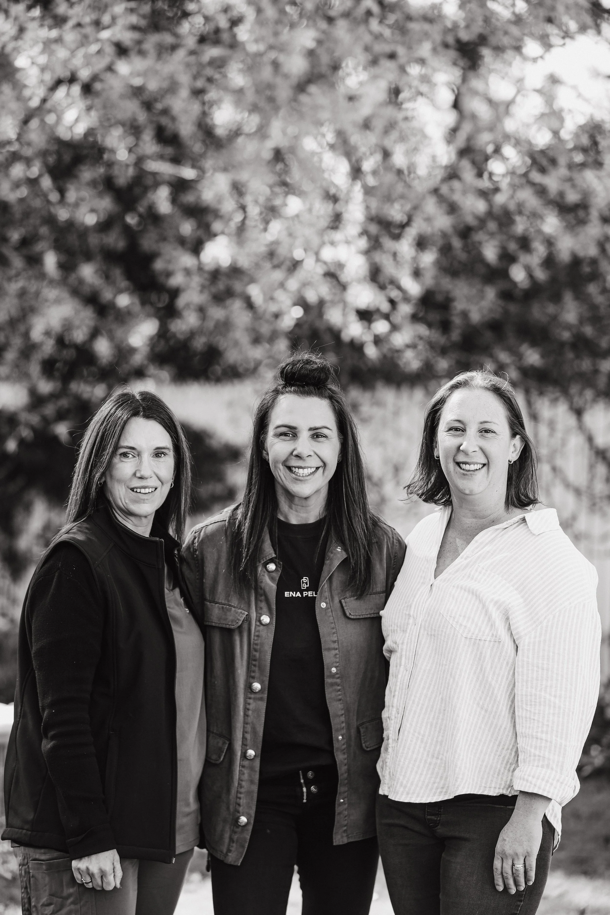 Three women standing outdoors in front of a blurred background of trees, smiling for a photo.