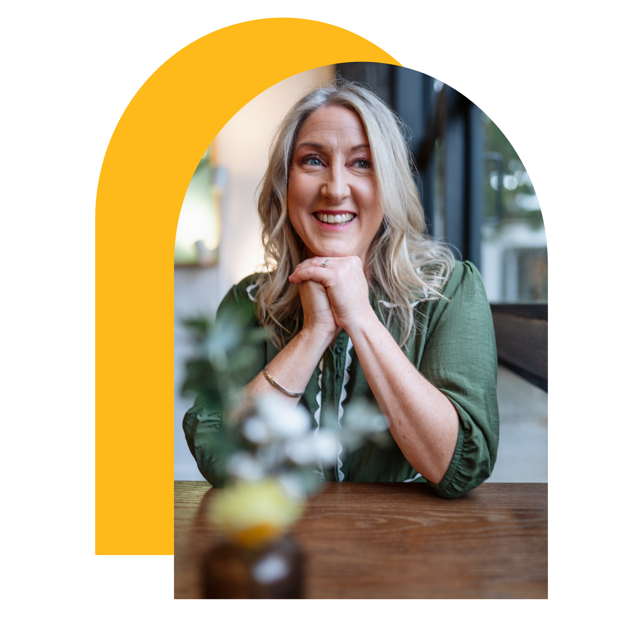 A woman with blonde curly hair smiling and resting her chin on her hand at a wooden table, with a blurred background of a window and some greenery, in a cozy indoor setting.