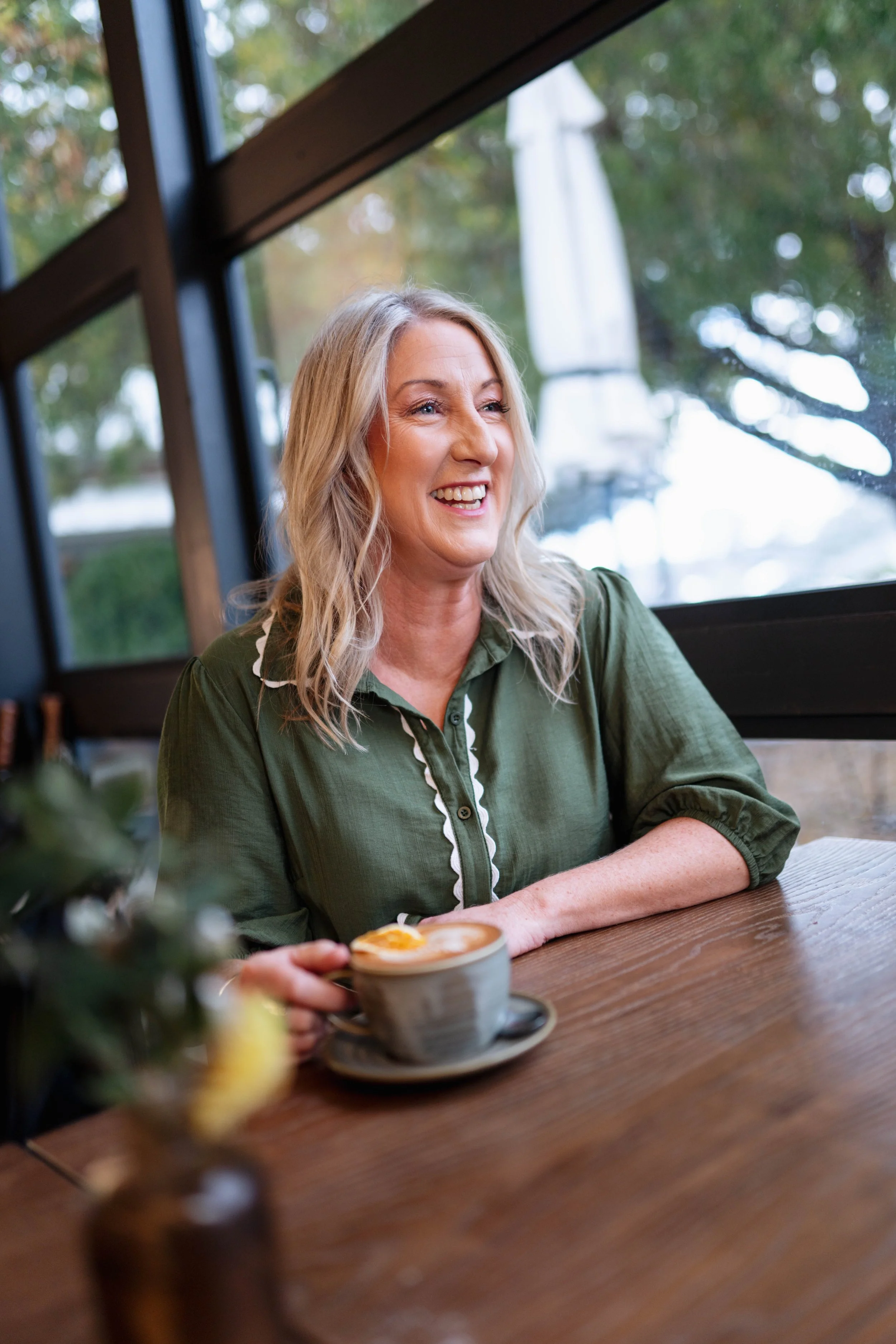 Mel white in a cafe setting, smiling and holding a cup of coffee