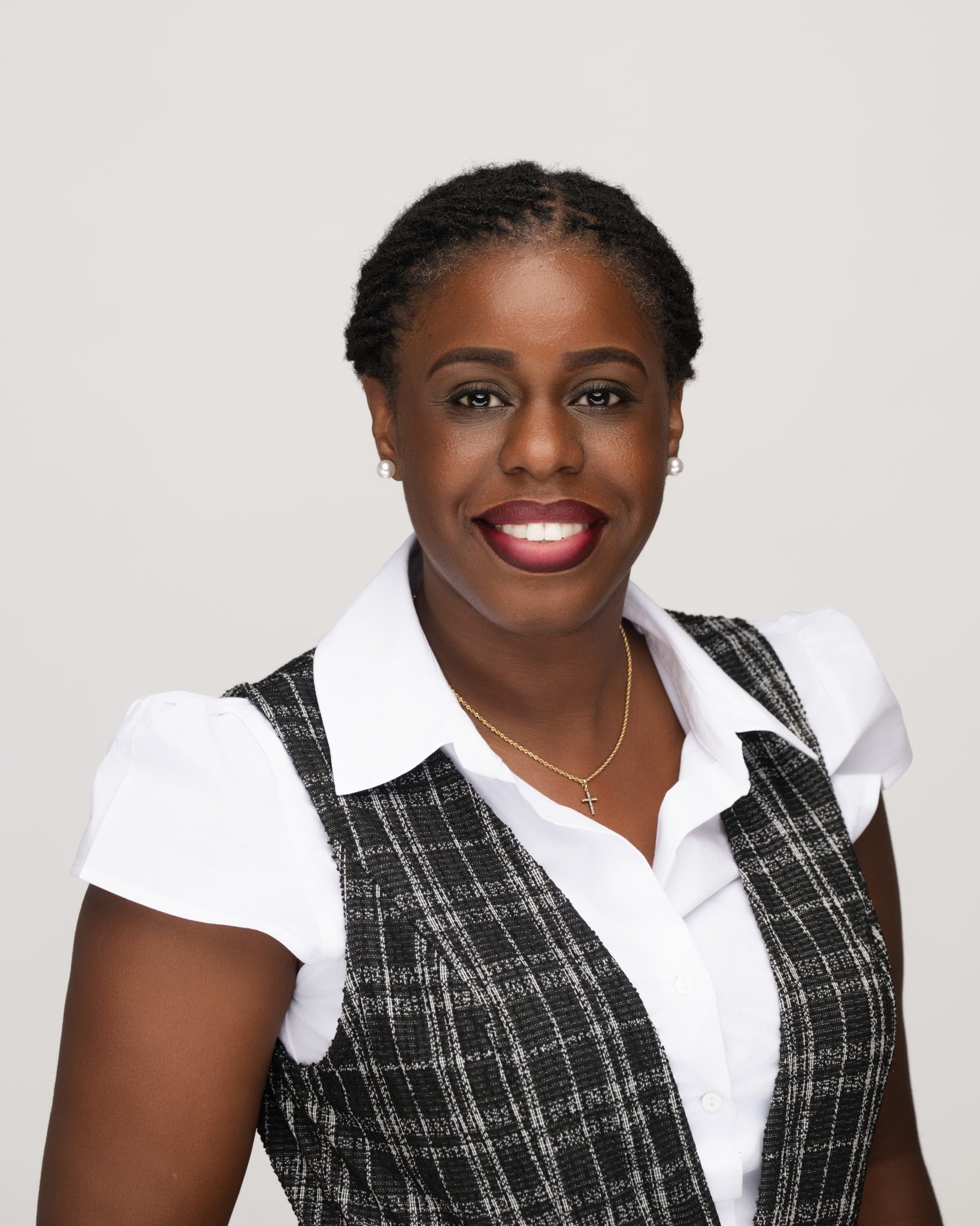 Close-up portrait of an African American woman smiling, wearing pearl earrings, a gold cross necklace, a white shirt, and a black and white plaid vest, against a plain light gray background.