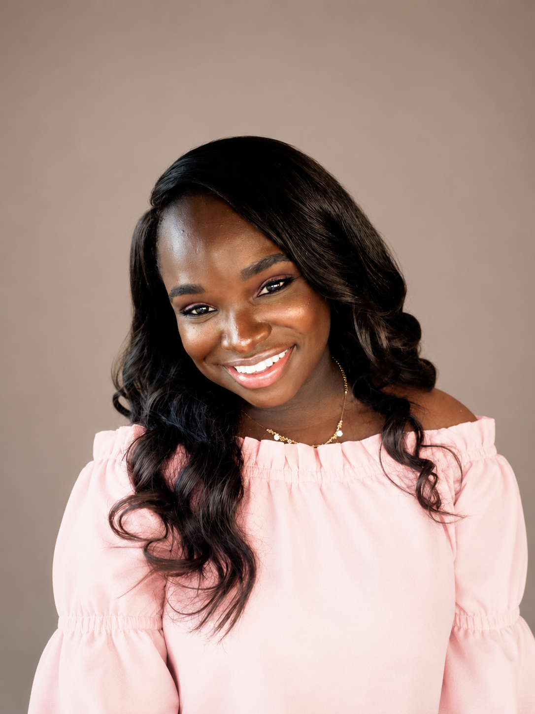 A smiling woman with dark hair wearing a pink off-shoulder top and a delicate gold necklace with flowers, against a plain neutral background.