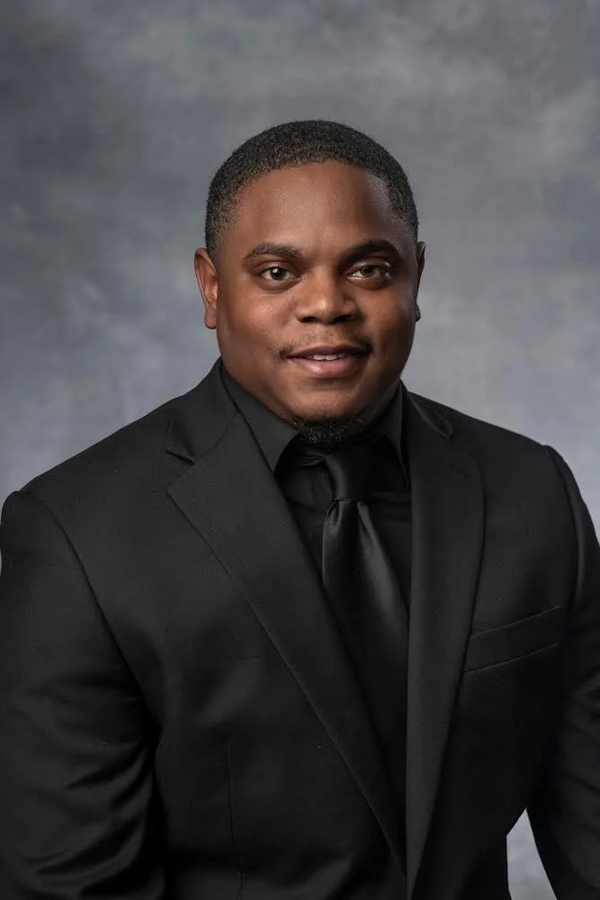 A young man in a black suit, black shirt, and black tie, smiling against a gray background.