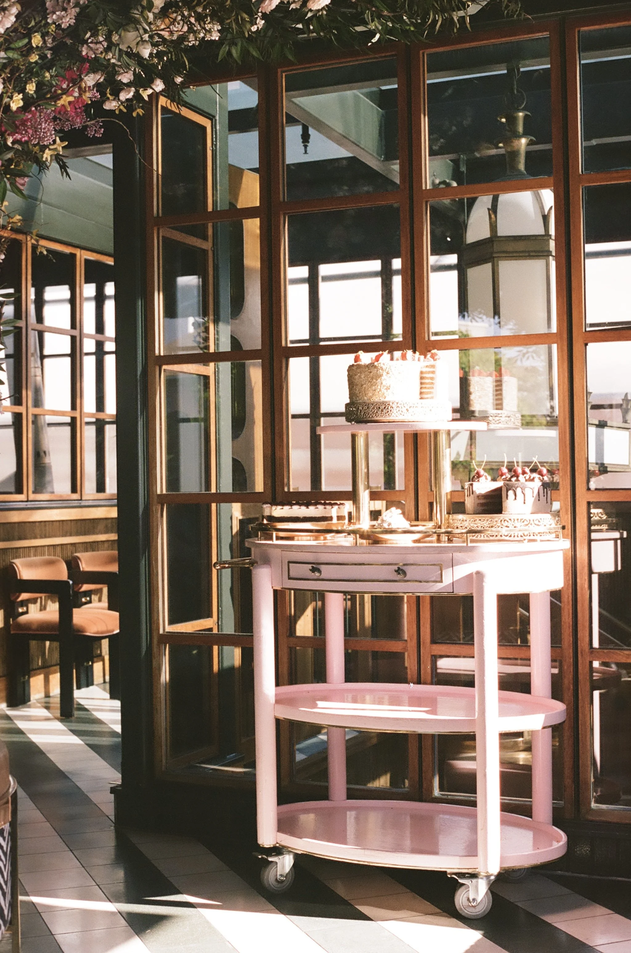A pink serving cart with two shelves and wheels, situated in a restaurant or cafe. Behind the cart is a large window with wooden frames, reflecting the interior and some decorative elements. The space is adorned with flowers and greenery, and there are chairs with orange cushions in the background.