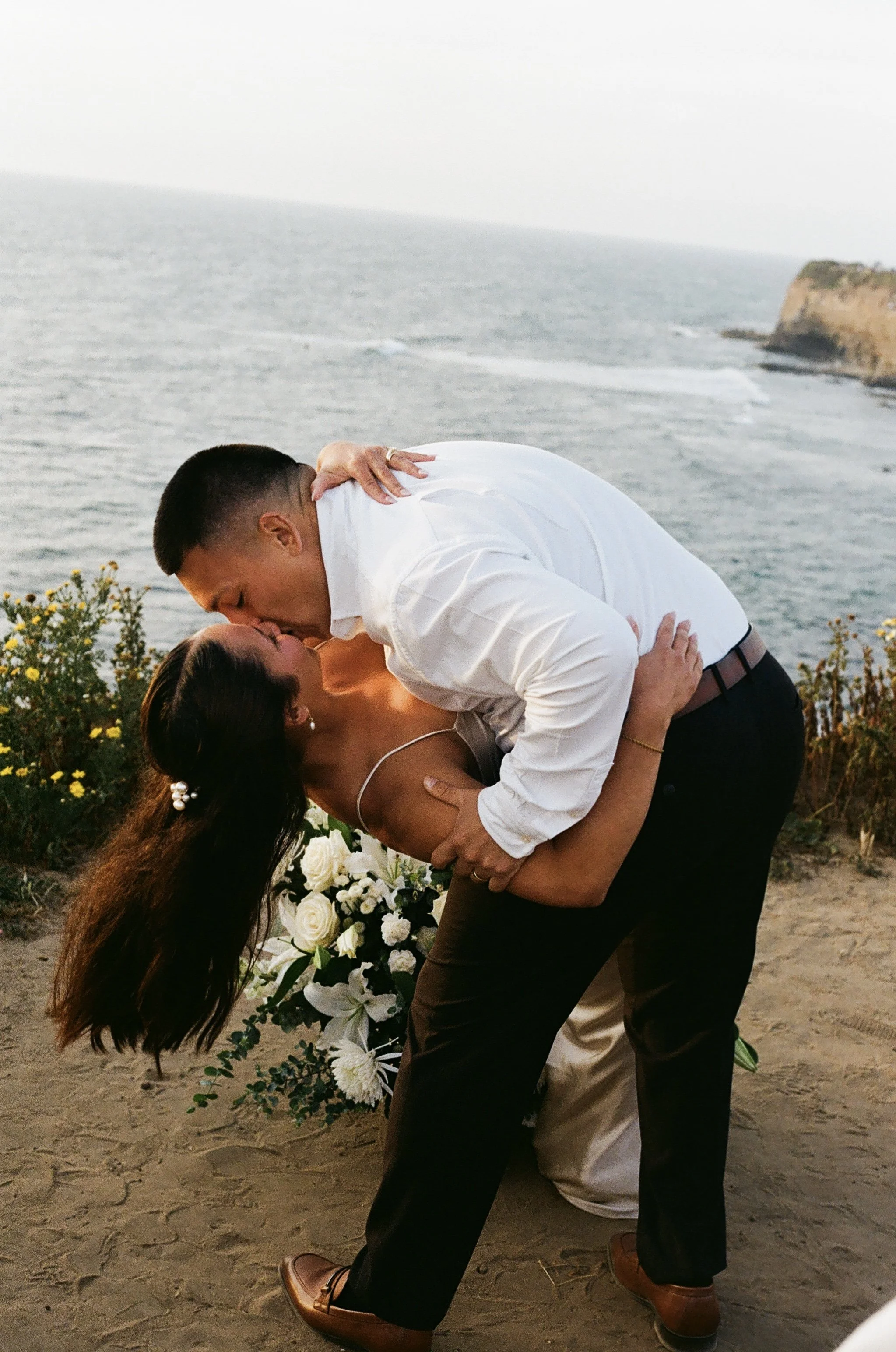 A couple sharing a kiss at a beachside wedding, with the ocean, a cliff, and part of a floral wedding decorations in the background.