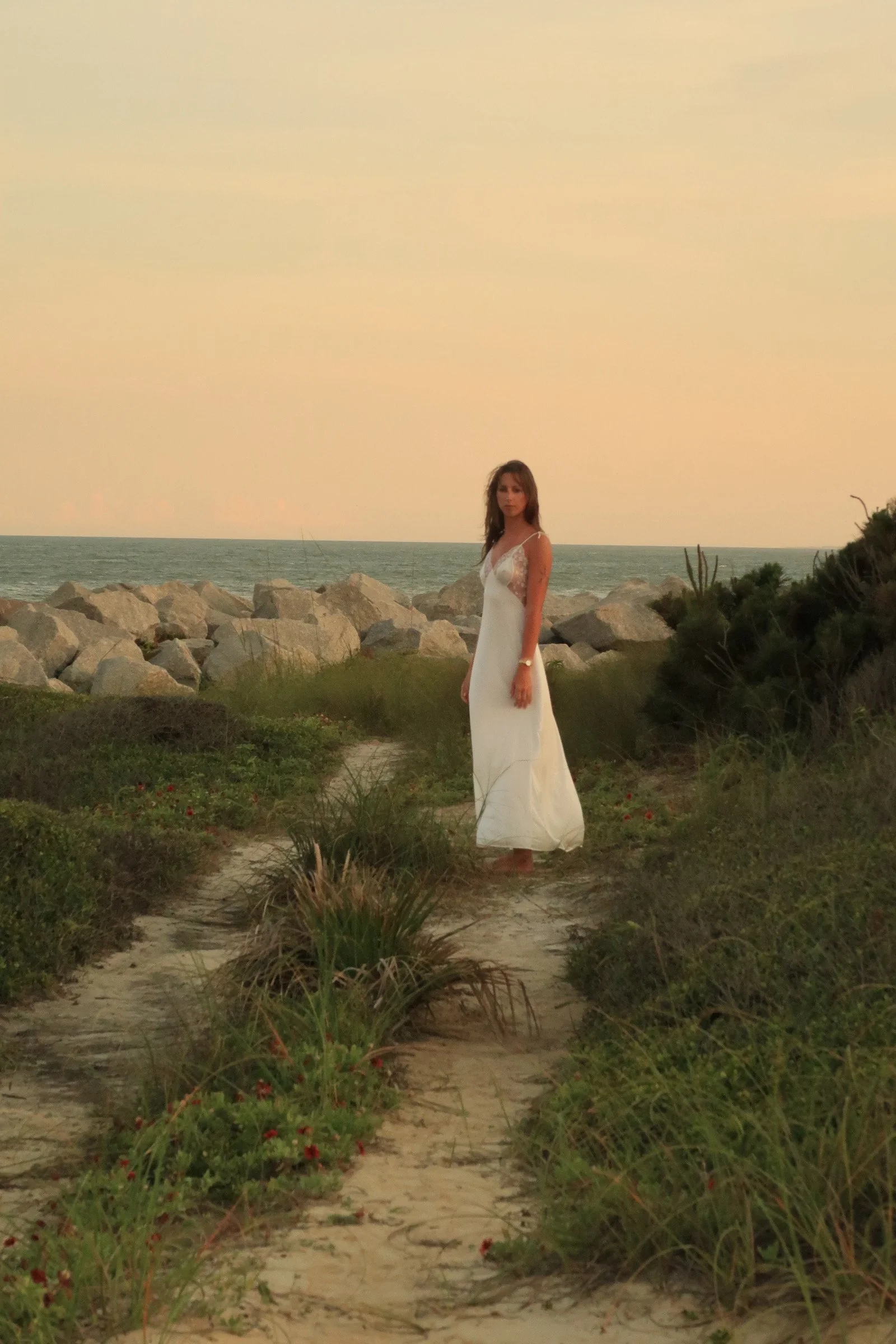 A woman in a white dress standing on a sandy path surrounded by green bushes and plants, with rocks and the ocean in the background during sunset or sunrise.