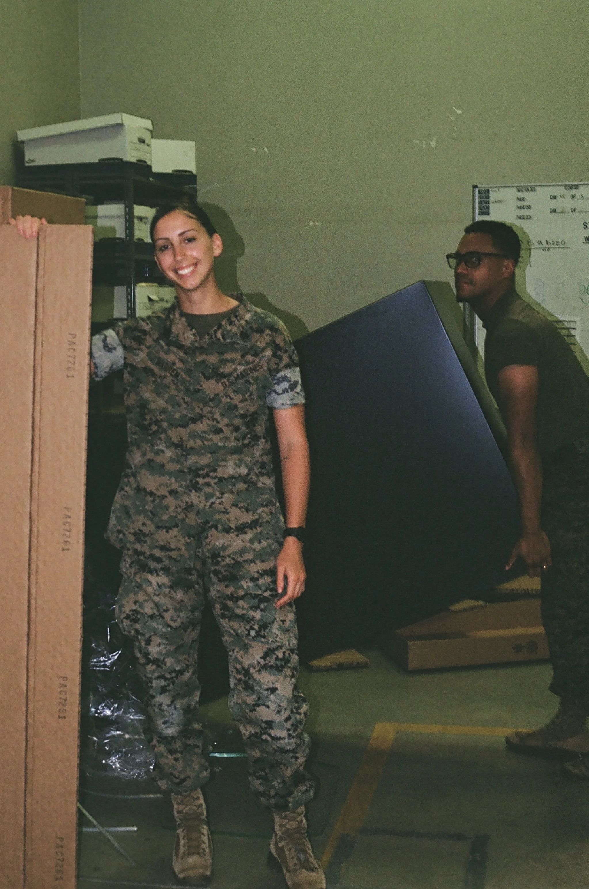 Two people, a woman and a man, are in a room with a whiteboard and boxes. The woman is smiling, wearing camouflage military uniform and standing near a cardboard box. The man is wearing glasses and dark clothing, standing behind a large computer monitor, looking to the side.