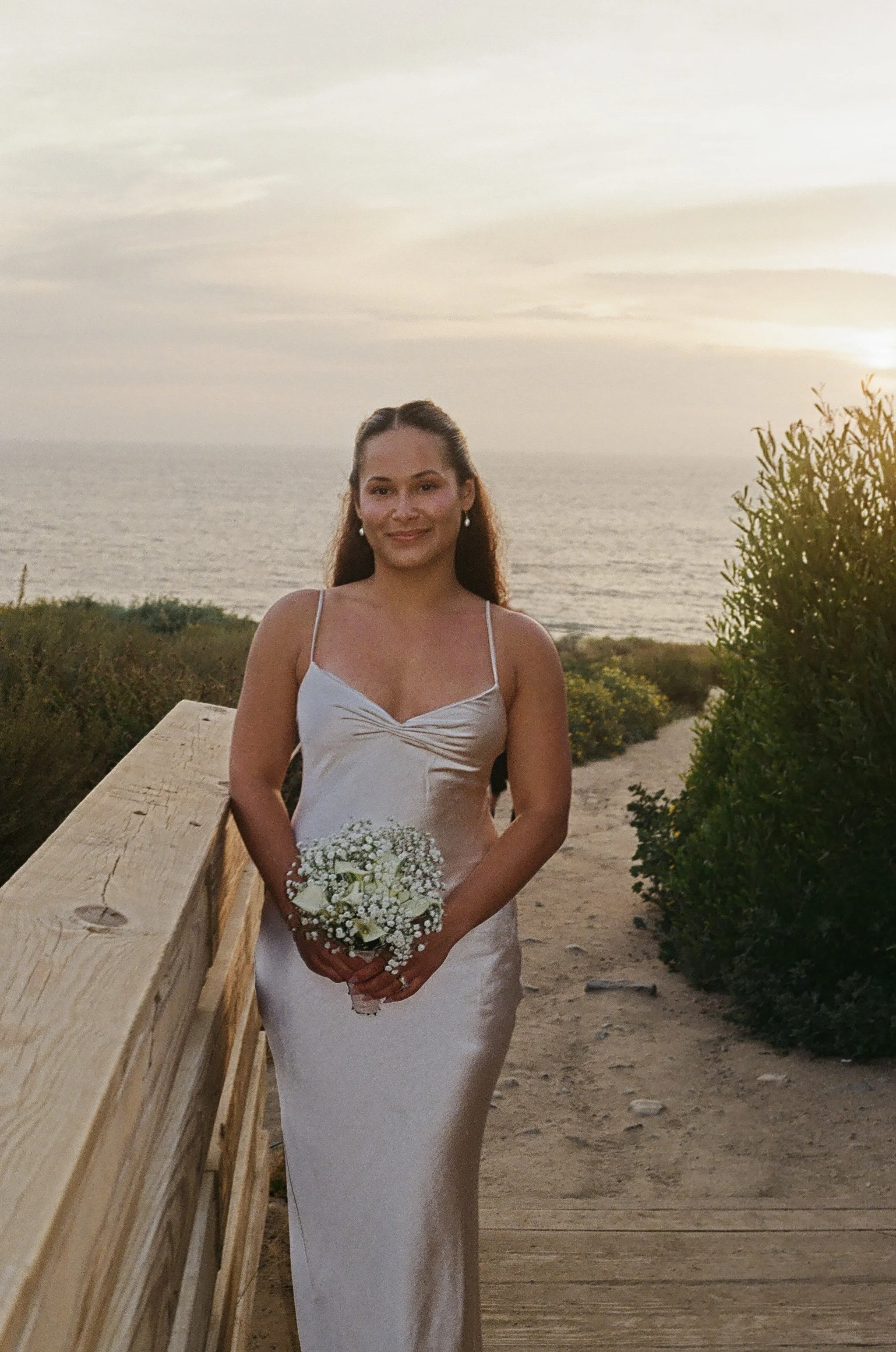 A woman in a white dress holding a bouquet of white flowers poses outdoors at sunset near the ocean, with greenery and a wooden railing in the background.