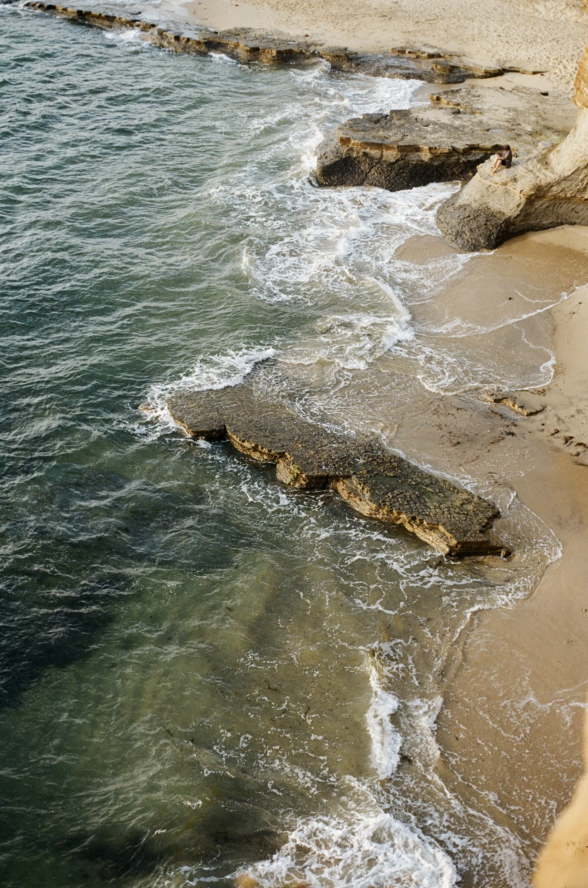 A view of the coastline showing waves hitting rocks and sandy beach with a person sitting on a large rock.