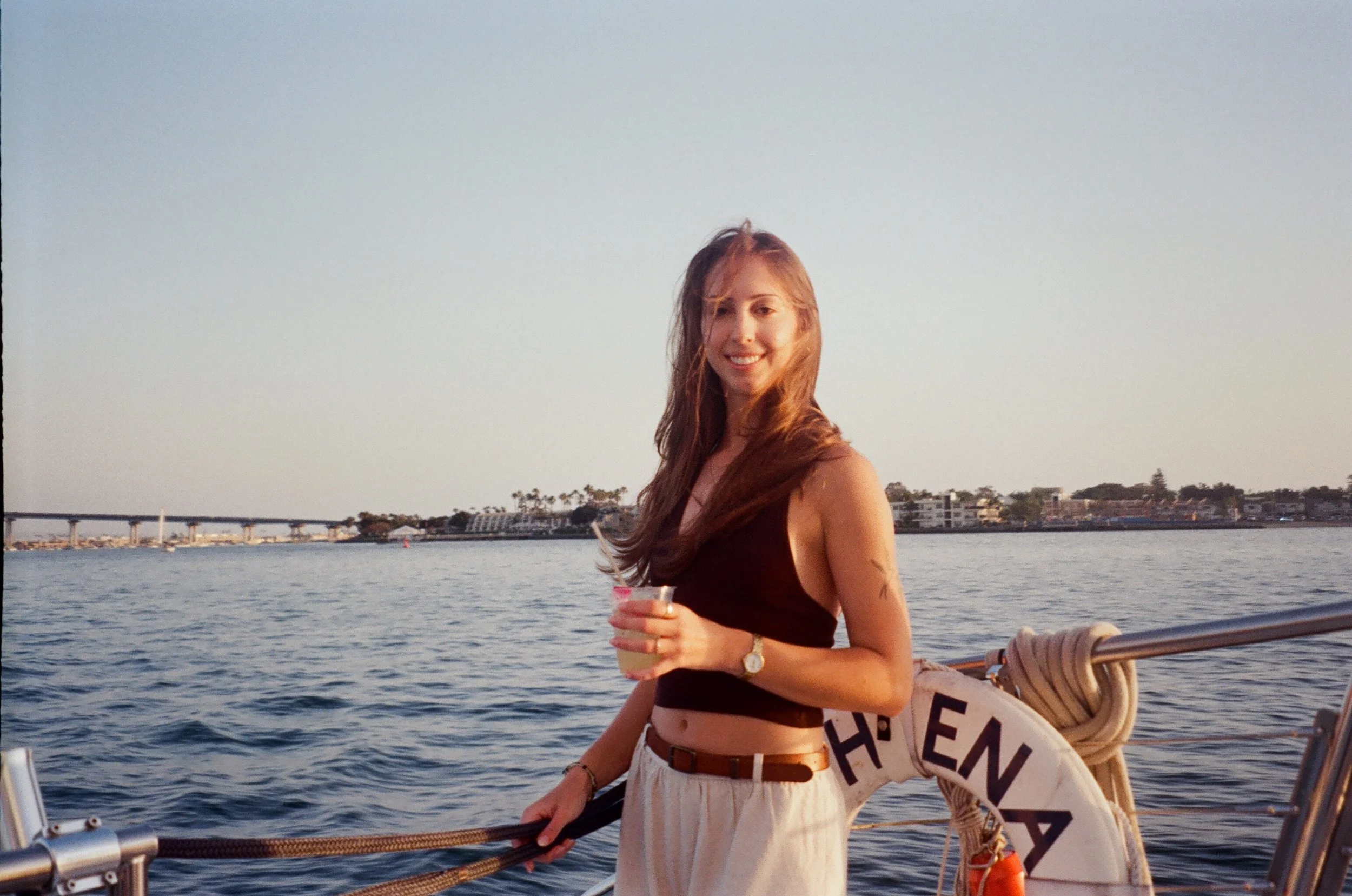 A young woman with long red hair smiling and holding a drink, standing on a boat near the water at sunset with a cityscape and bridge in the background.