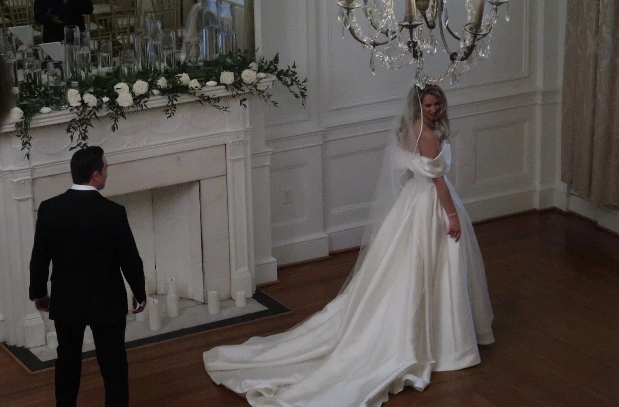 A bride in a white wedding dress with a long train and veil is smiling while looking over her shoulder during her wedding. A groom in a black tuxedo is standing nearby in a decorated indoor venue with candles on the floor in front of a fireplace, floral arrangements, and a crystal chandelier hanging from the ceiling.