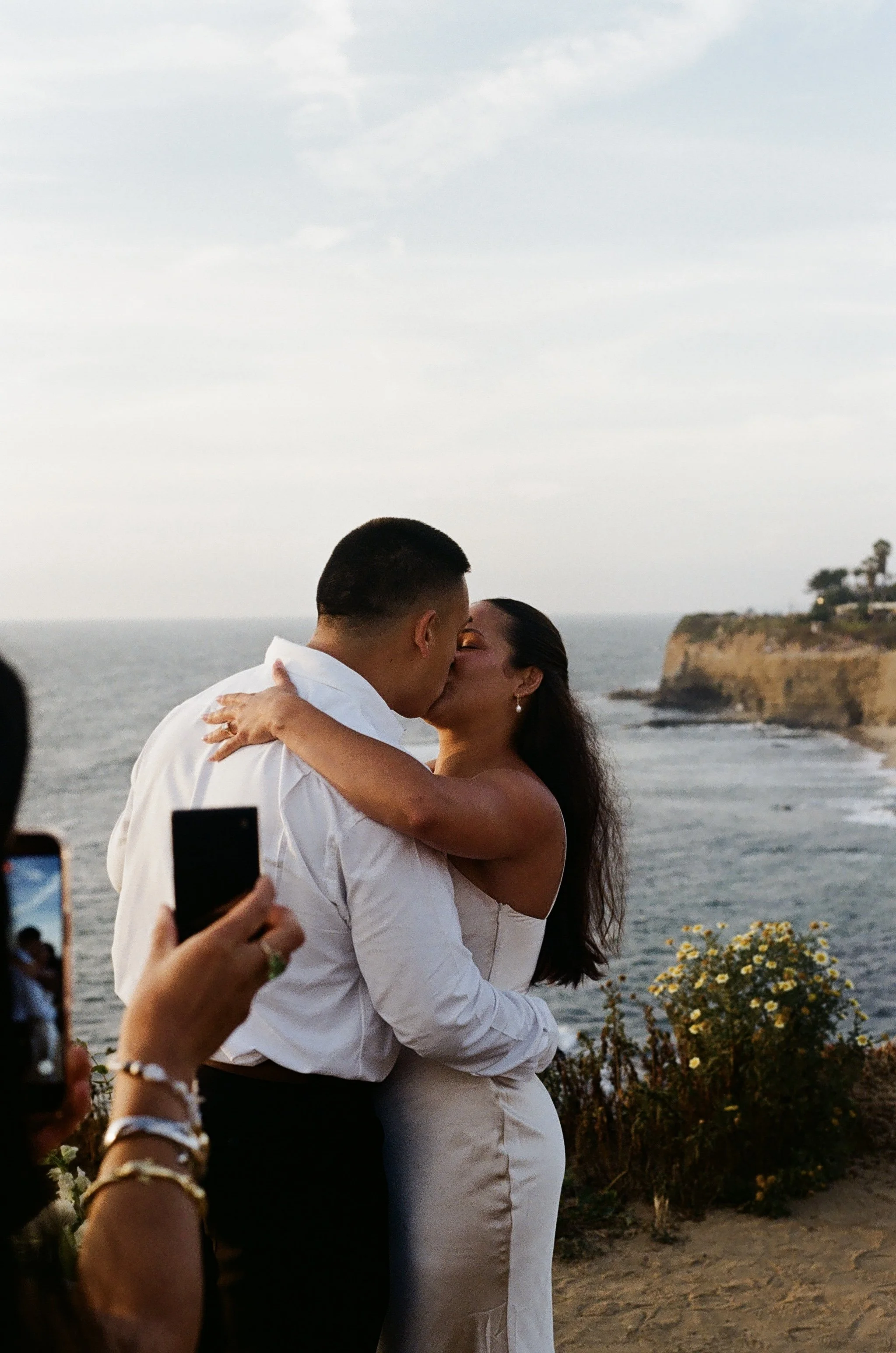 A couple sharing a kiss at sunset on a beach, dressed in wedding attire, with people taking photos around them and a scenic coastline in the background.