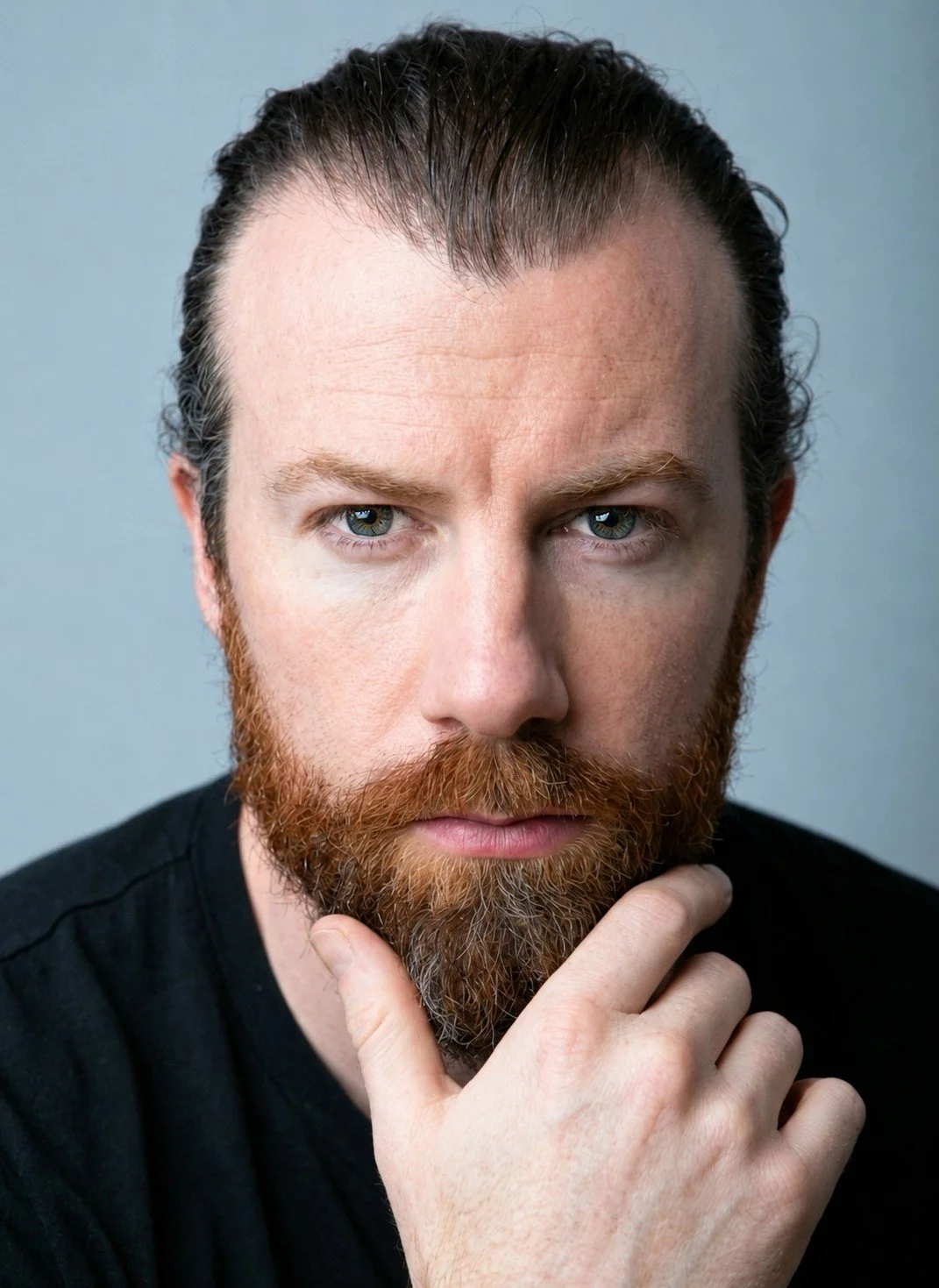 Close-up of a man with red beard and blue eyes, holding his chin with his hand, wearing a black shirt, against a plain light background.