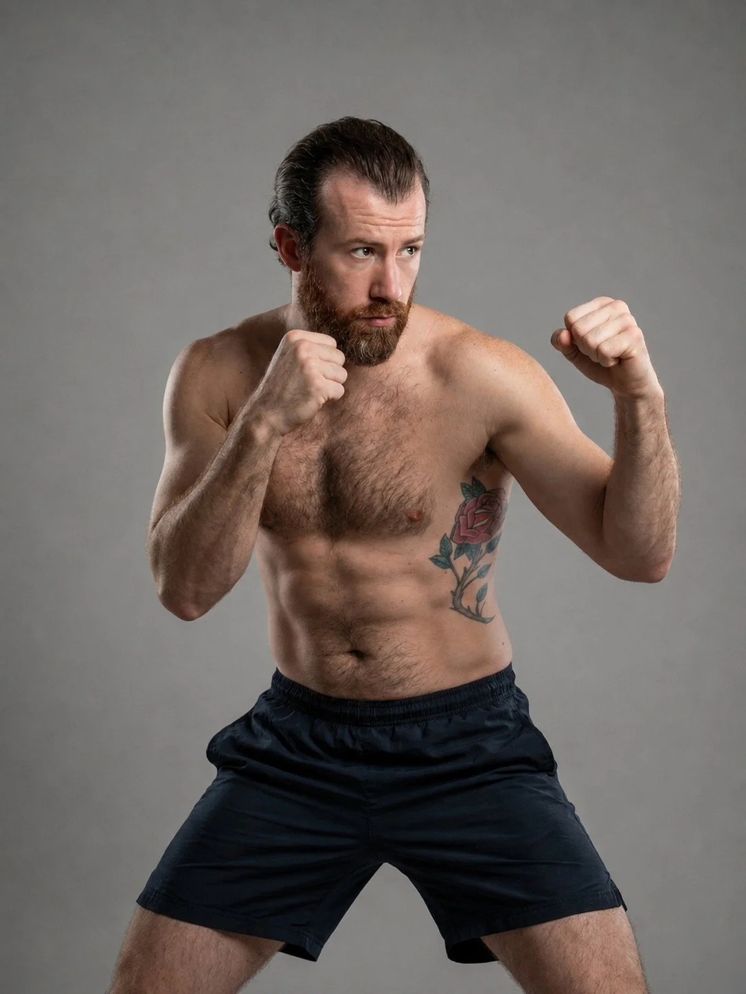 A shirtless man with a beard and a tattoo of a rose on his left side, standing in a martial arts pose with fists clenched against a plain gray background.