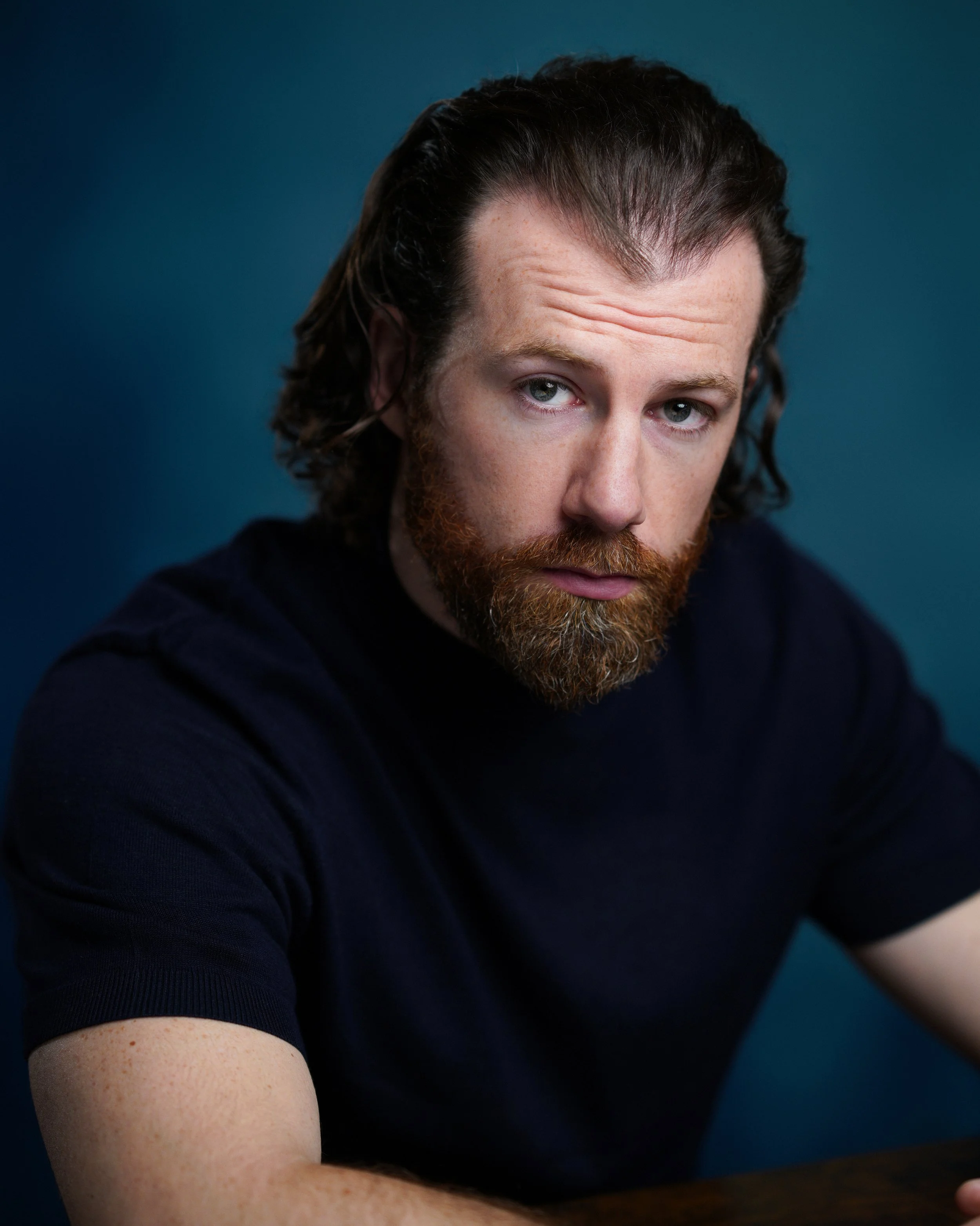 Close-up of a man with dark hair, a full beard, and light skin, wearing a dark shirt, against a dark blue background, looking at the camera.
