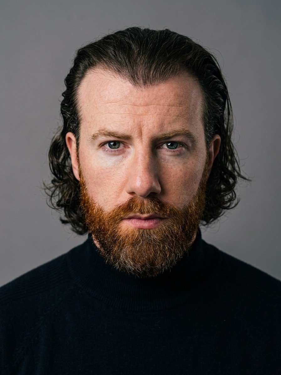 A man with long dark hair and a full red beard wearing a black turtleneck, looking directly at the camera with a serious expression.