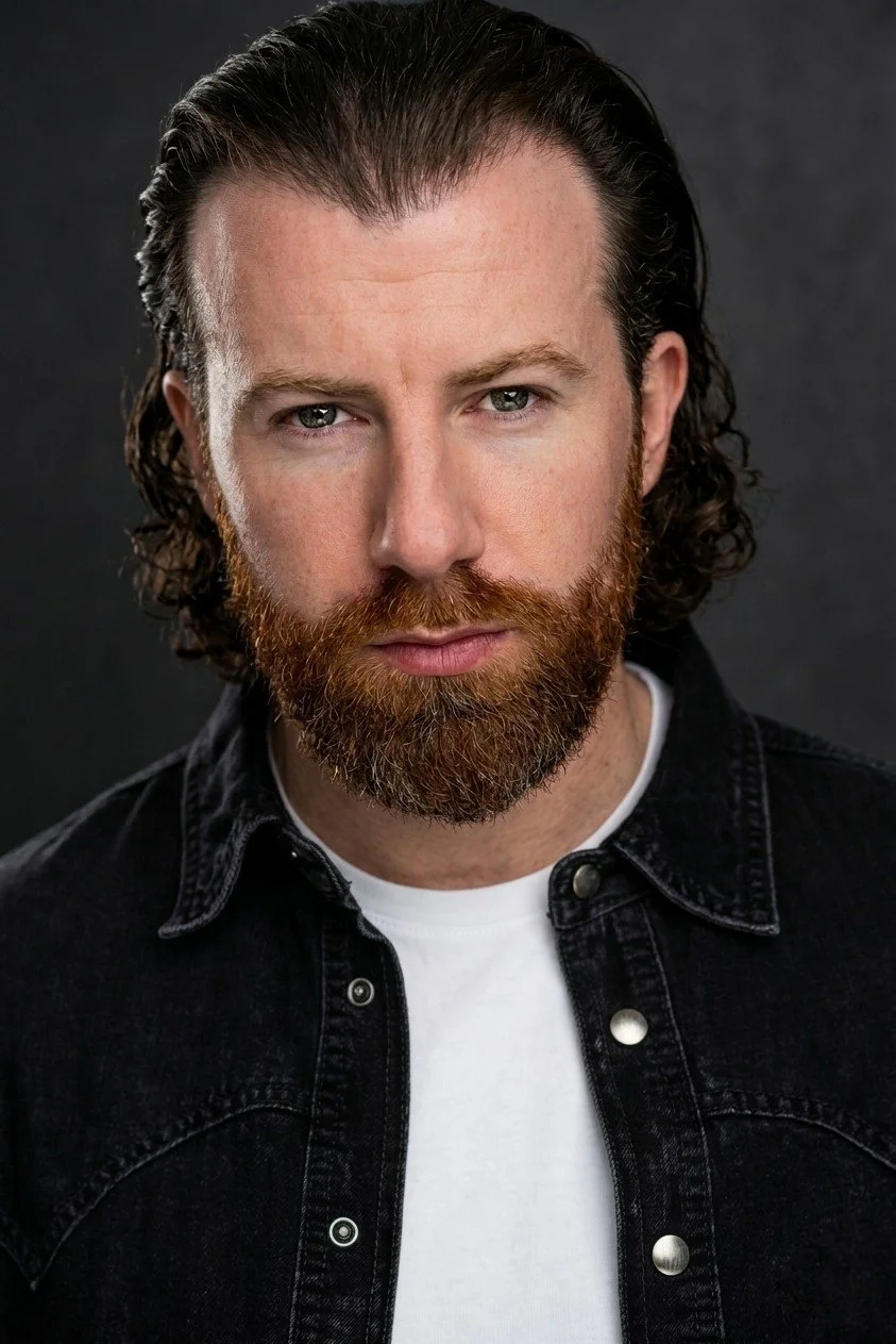 Close-up portrait of a man with long, dark, wavy hair, a full red beard, and intense blue eyes, wearing a black denim jacket over a white t-shirt against a dark background.