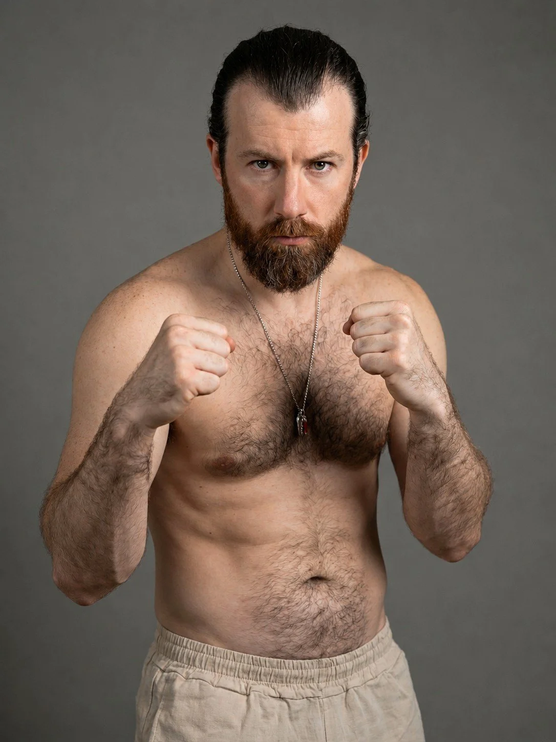 A shirtless man with a beard and slicked-back hair, wearing a chain necklace, poses with fists raised in a fighting stance against a plain gray background.