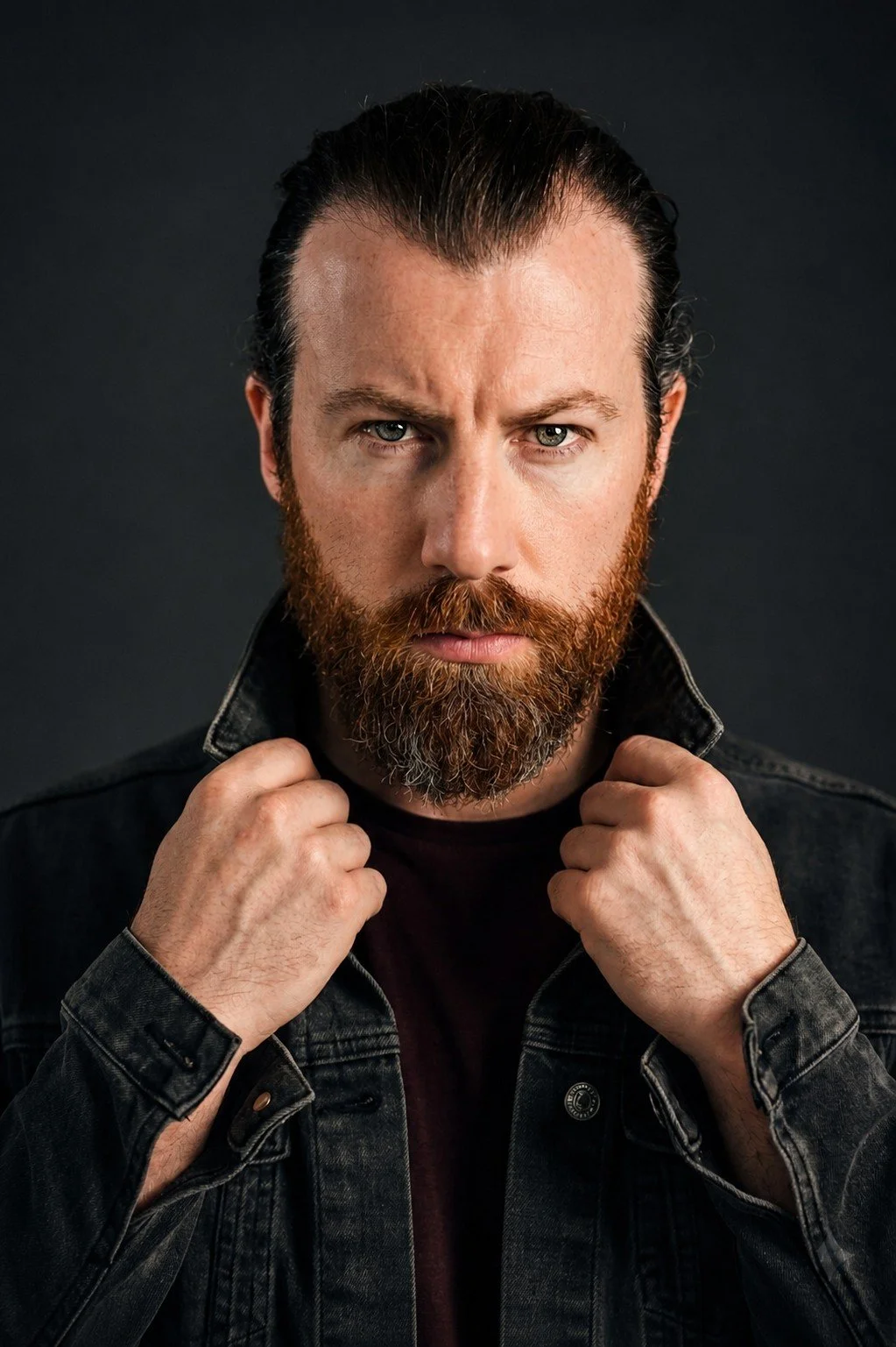 A man with a serious expression, beard, and slicked-back hair, holding the collar of his black denim jacket against a dark background.