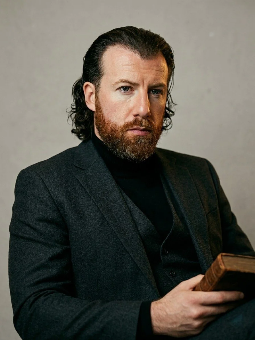 A man with shoulder-length dark hair and a beard, wearing a dark blazer and black shirt, sitting against a plain gray background, holding a book in his hand, looking intently at the camera.
