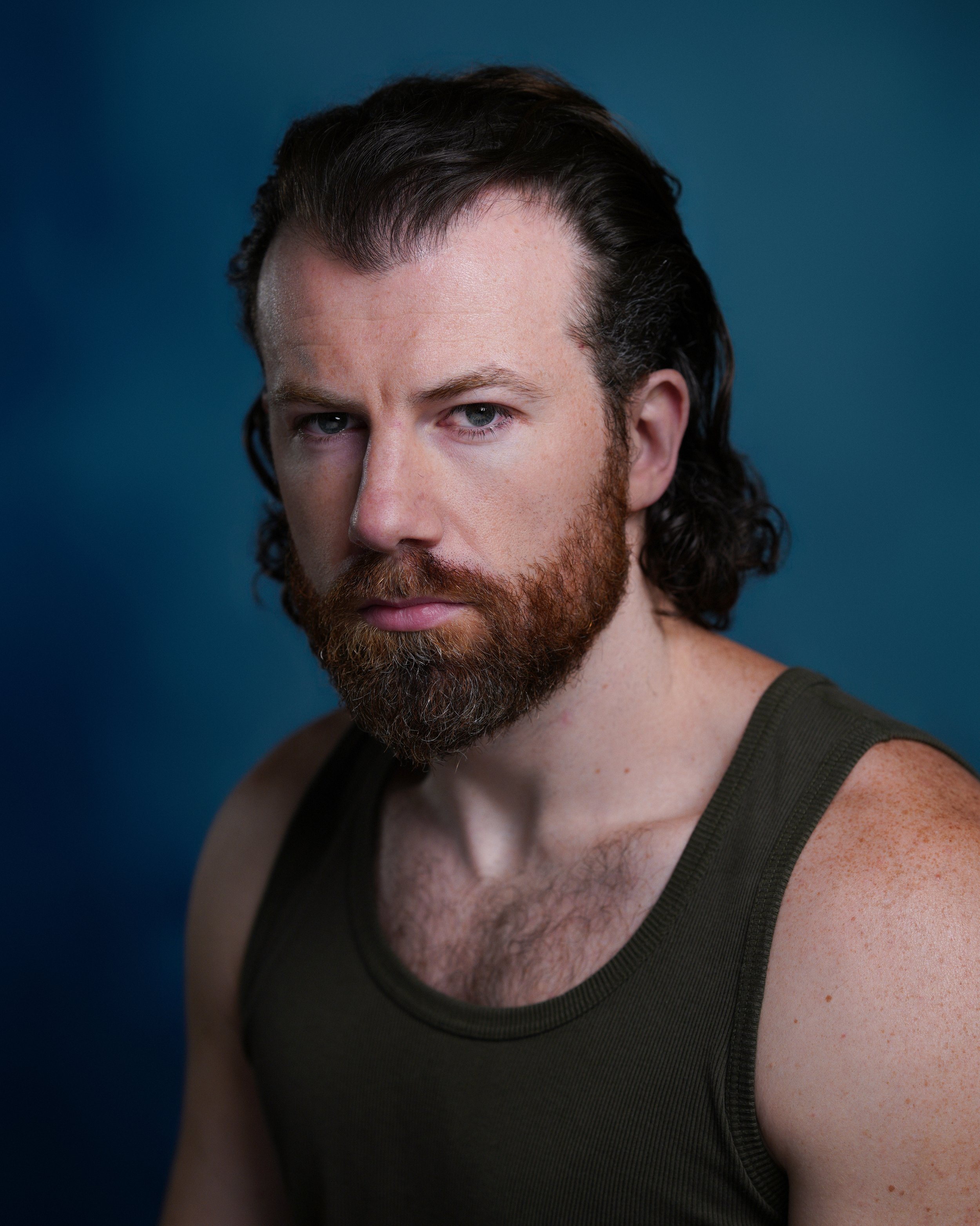 Close-up portrait of a man with long dark hair, a full beard, and wearing a green tank top, against a blue background.